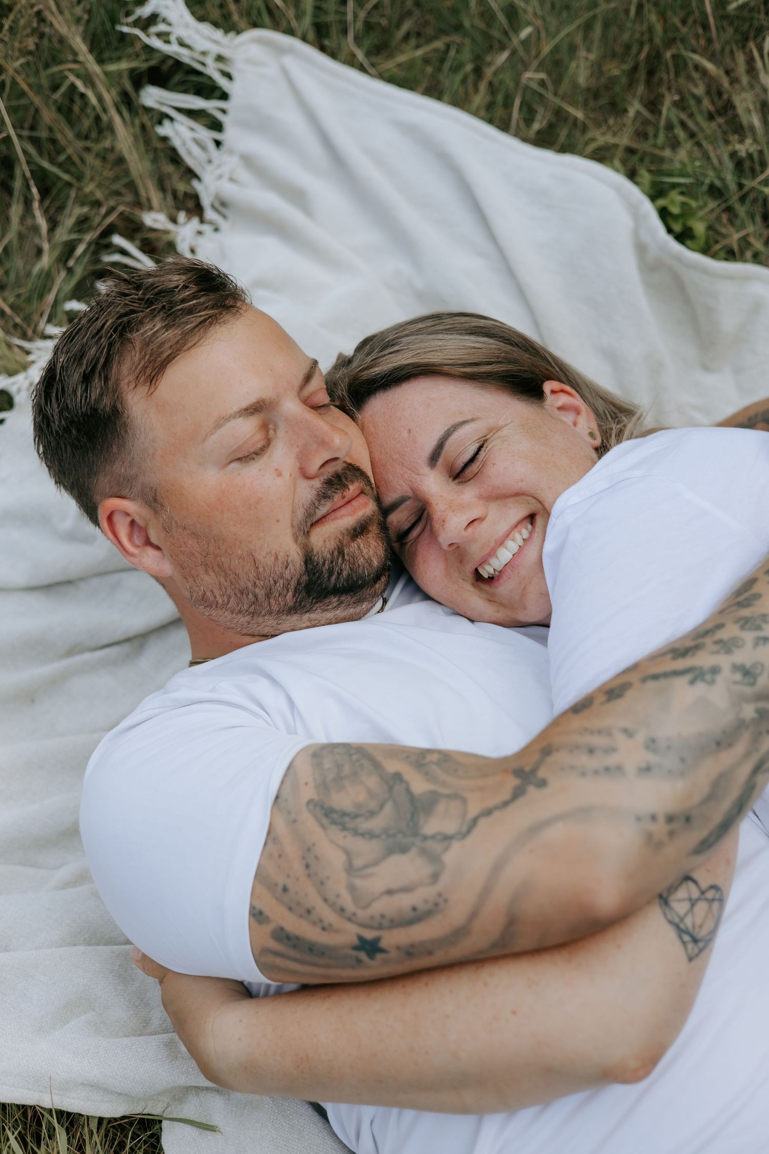 Couple cuddling on a blanket in a field, man with tattoos smiling.