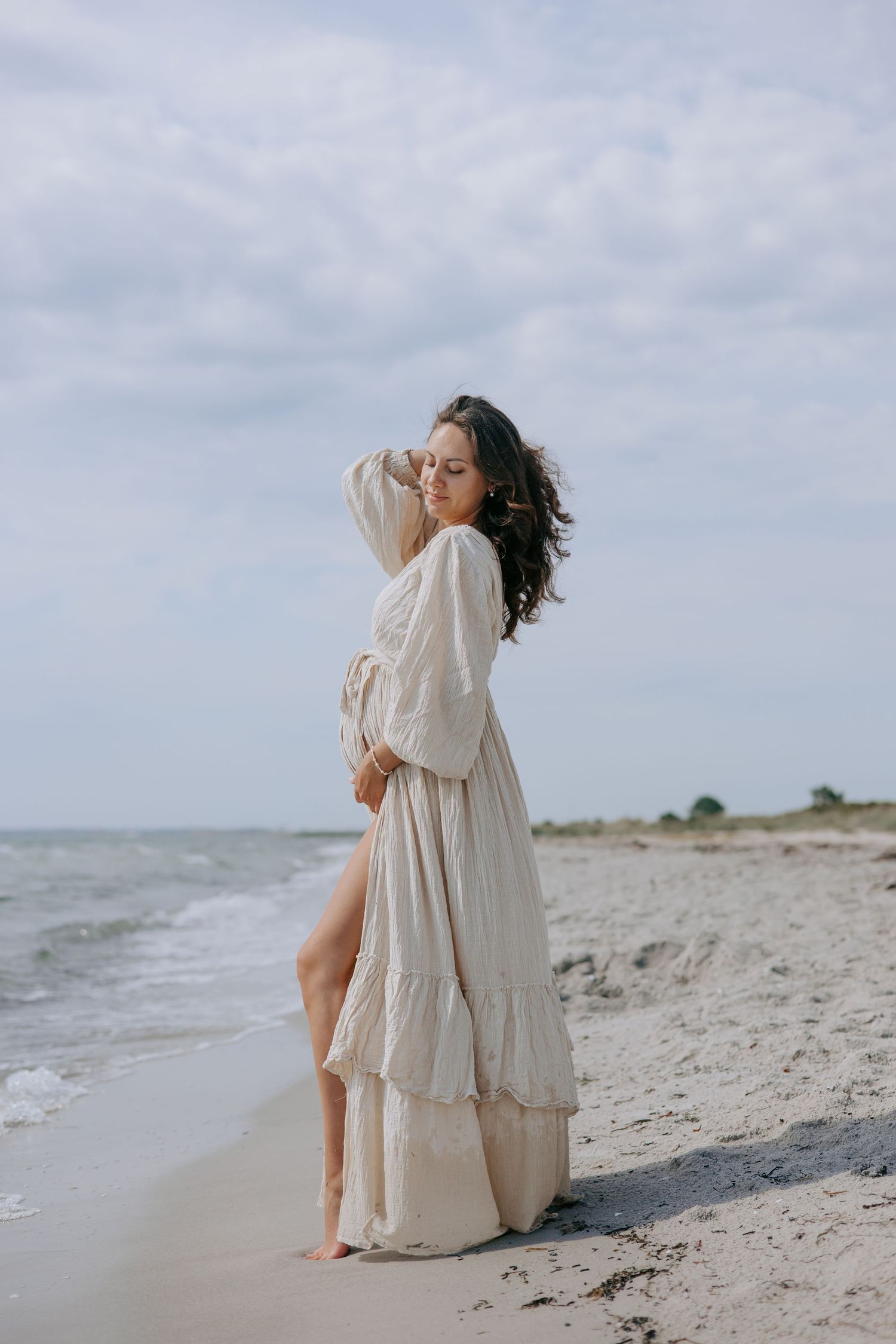 Pregnant woman in elegant cream ruffled dress posing on sandy beach with waves behind.