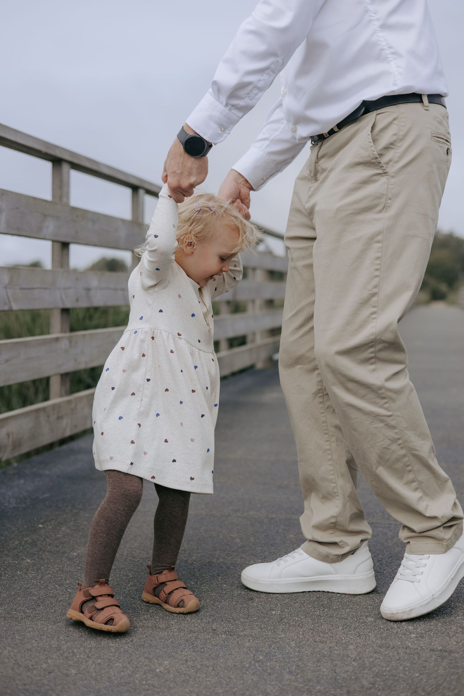 A blonde toddler girl in a white dress holding her fathers hand on a wooden bridge.