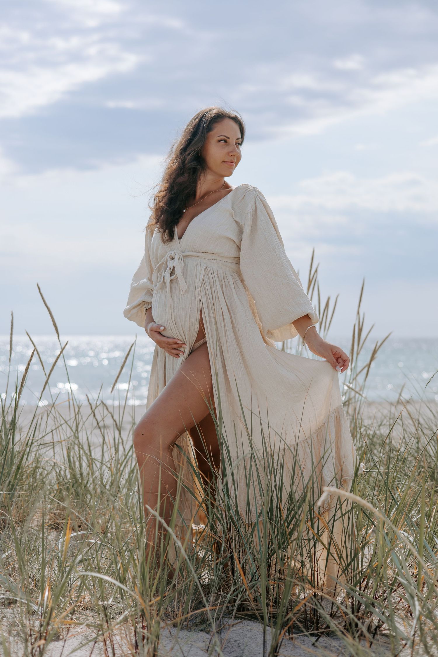 Pregnant woman in open beige linen dress standing in beach grass dunes with sparkling ocean behind.