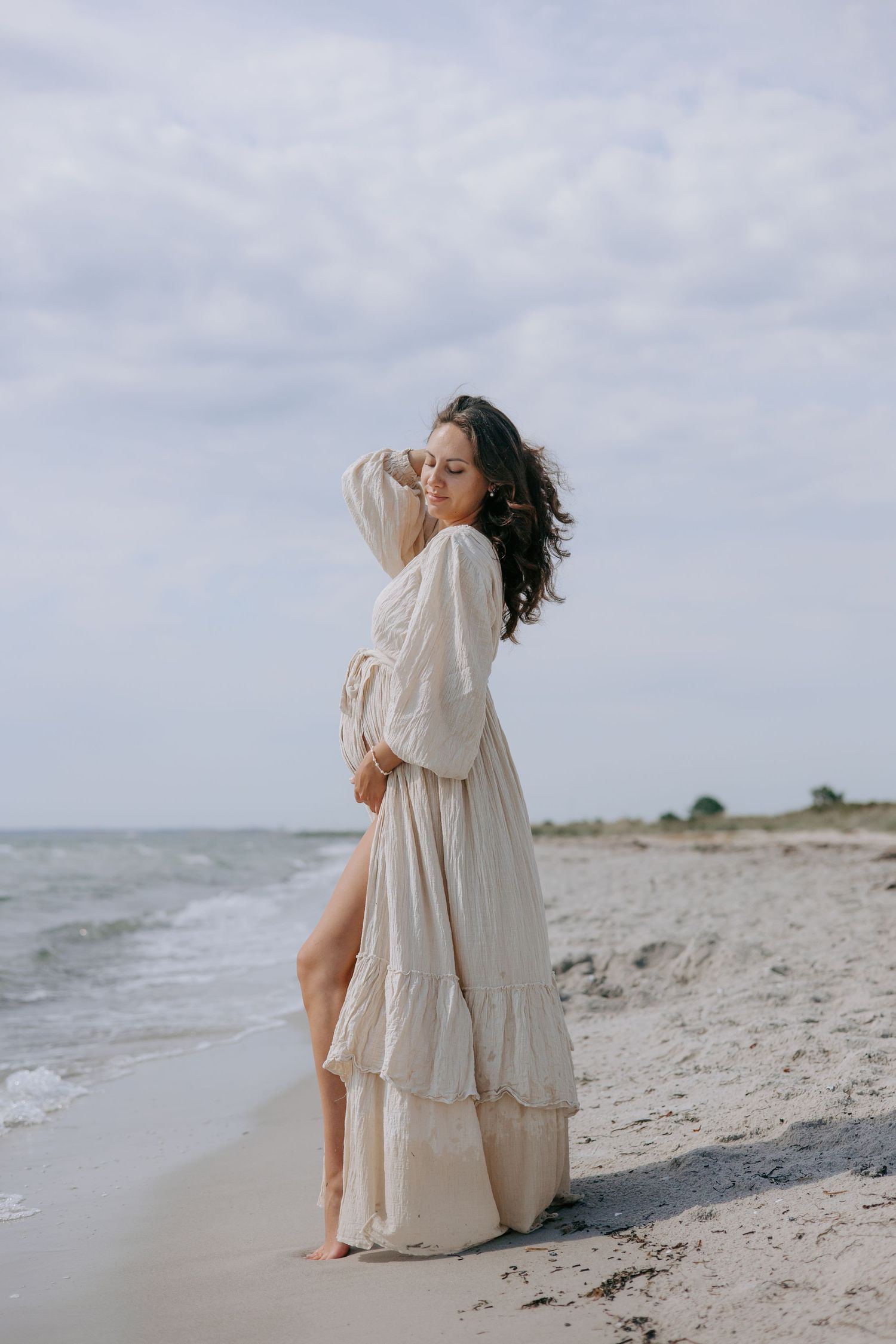 Pregnant woman in flowing beige maxi dress standing barefoot on sandy beach.