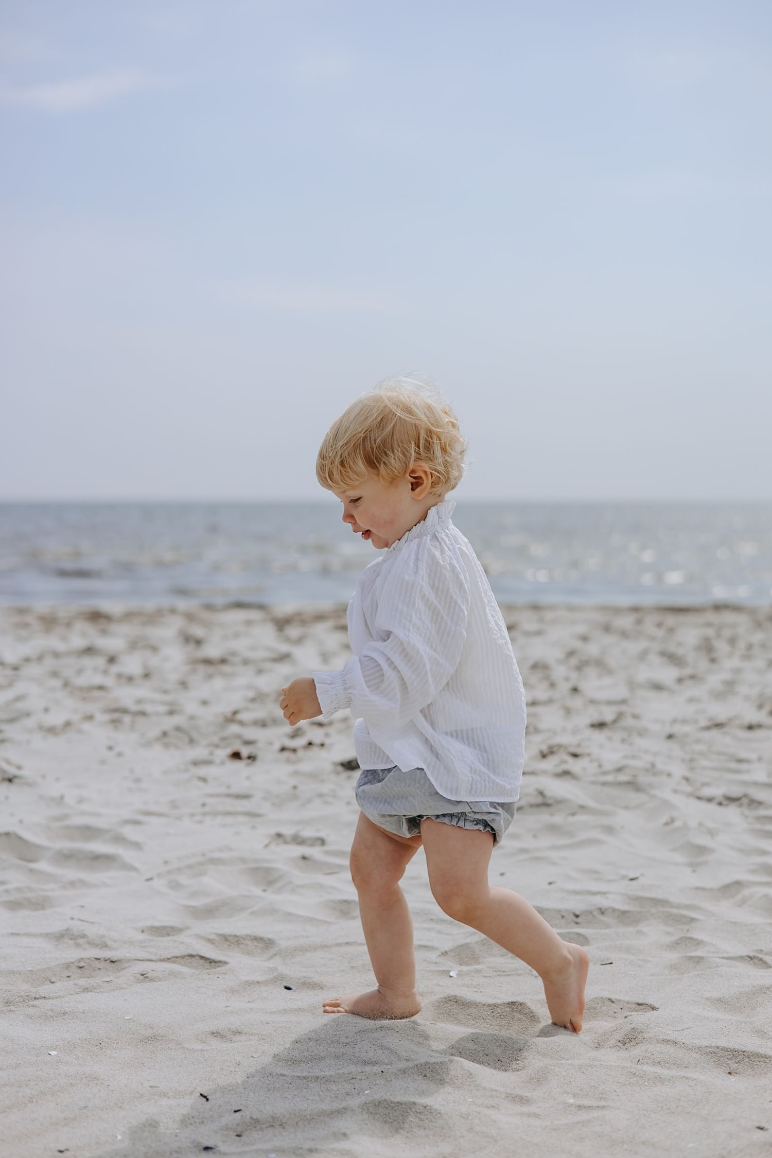 Blonde toddler in white shirt and denim shorts walking barefoot on sandy beach.