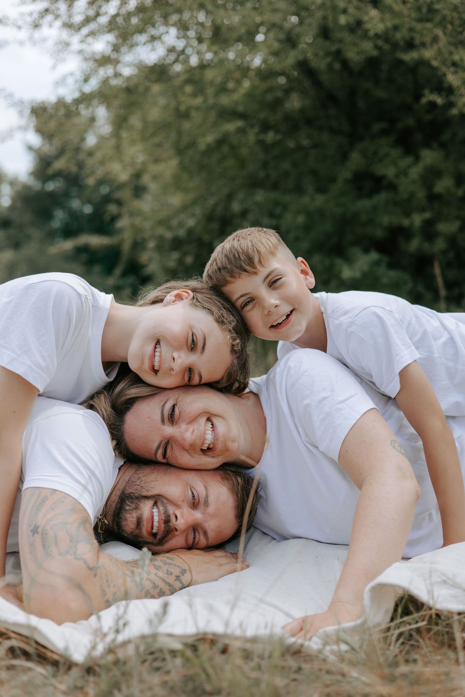 Happy family of four piled together on a blanket outdoors, smiling and laughing in white t-shirts.