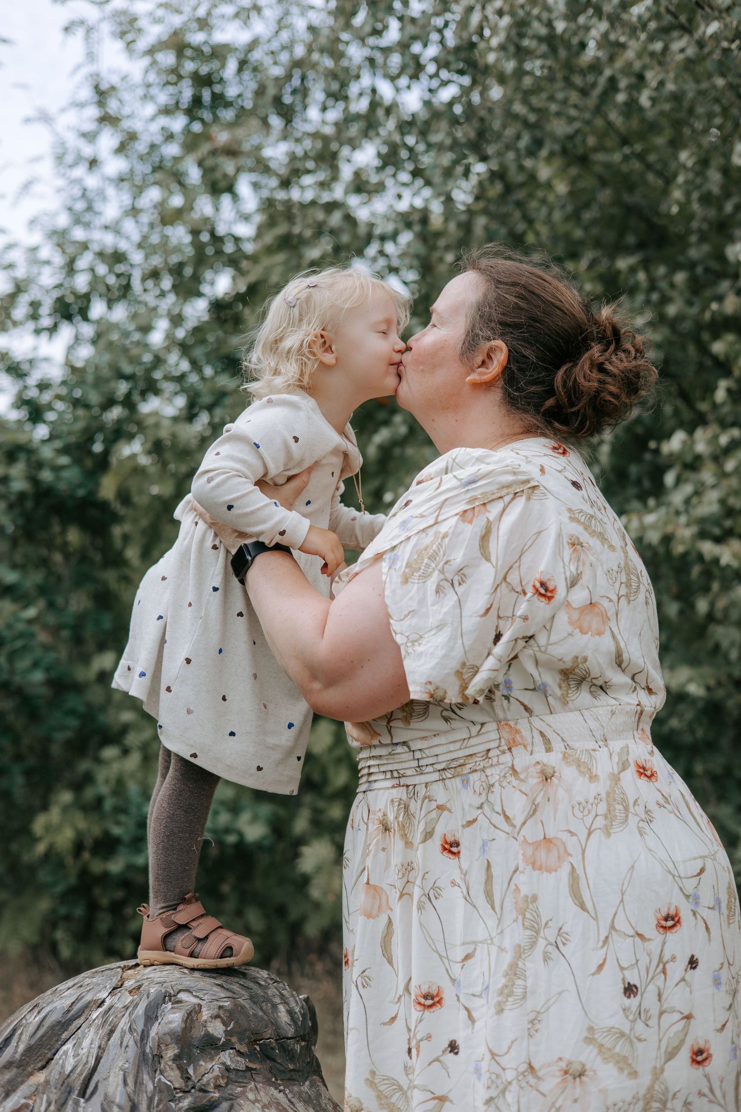 Mother in floral dress kissing young blonde toddler girl standing on rock outdoors.