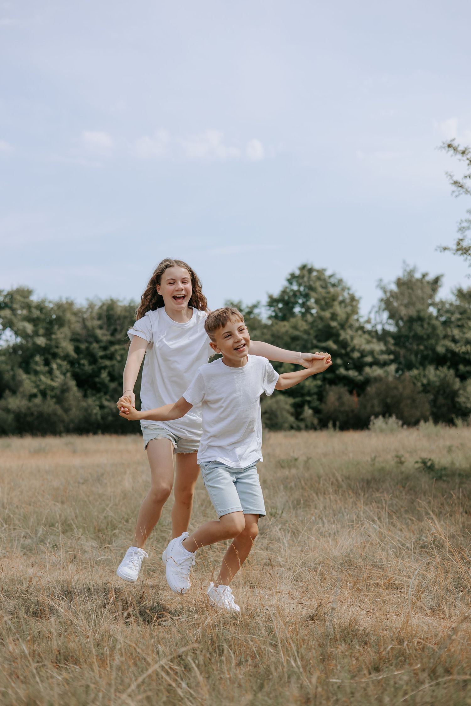 Two children in white t-shirts and blue shorts laughing and running together in an open grassy field.
