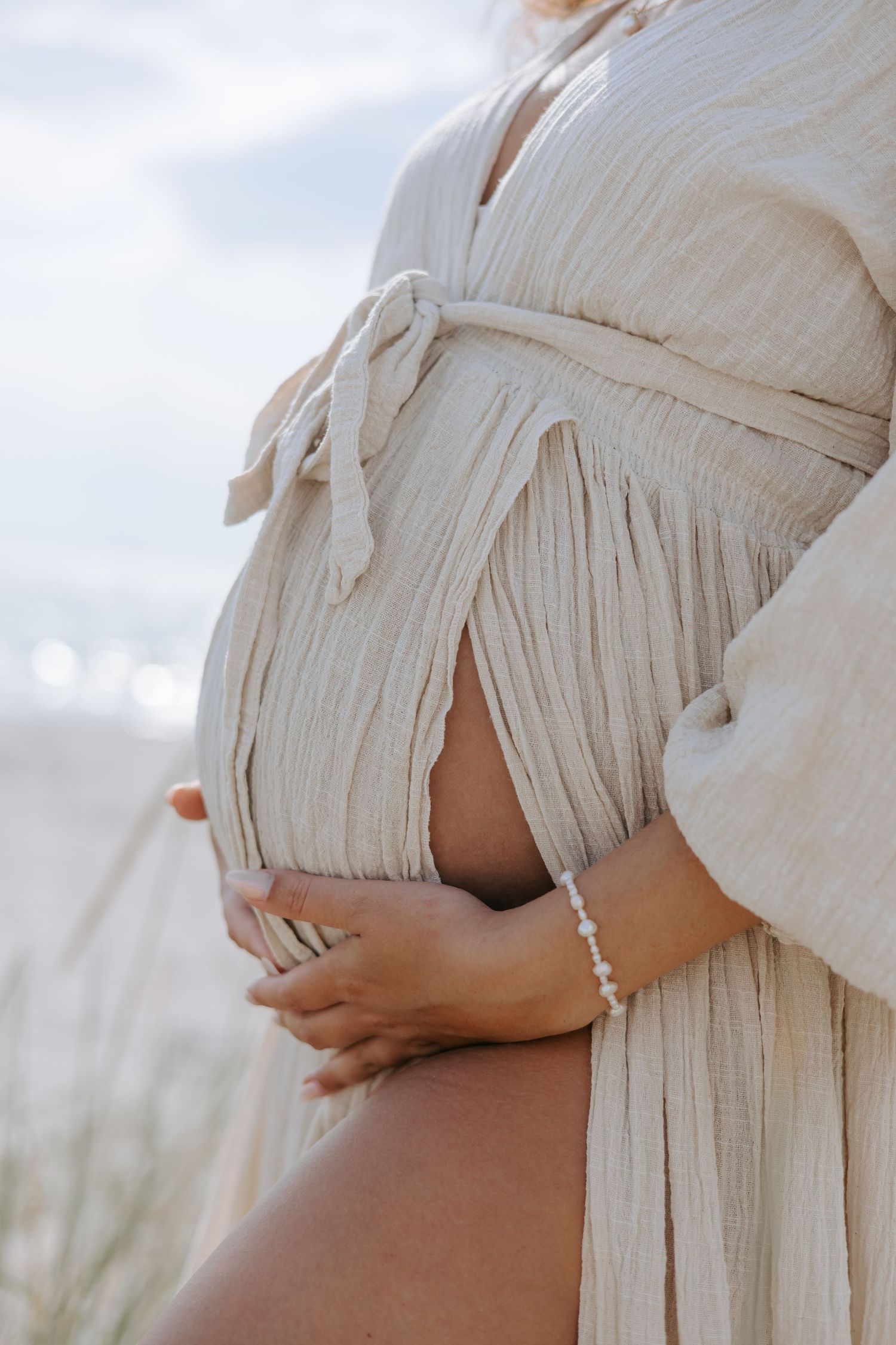 Close-up of a pregnant woman in a flowing beige linen dress cradling her baby bump at the beach.