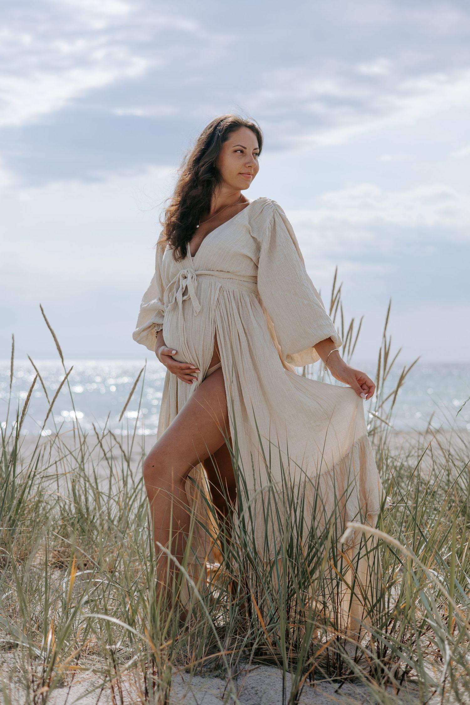Pregnant woman in a beige wrap dress standing among tall beach grasses with the ocean in the background.