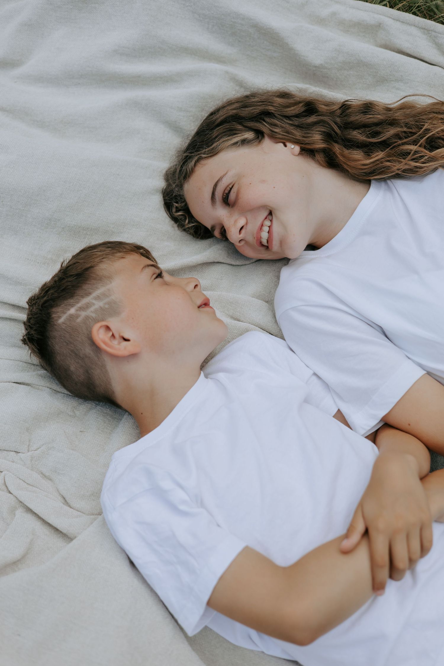 A smiling teenage girl and boy lying on a blanket outdoors, wearing white t-shirts, sharing a joyful moment.