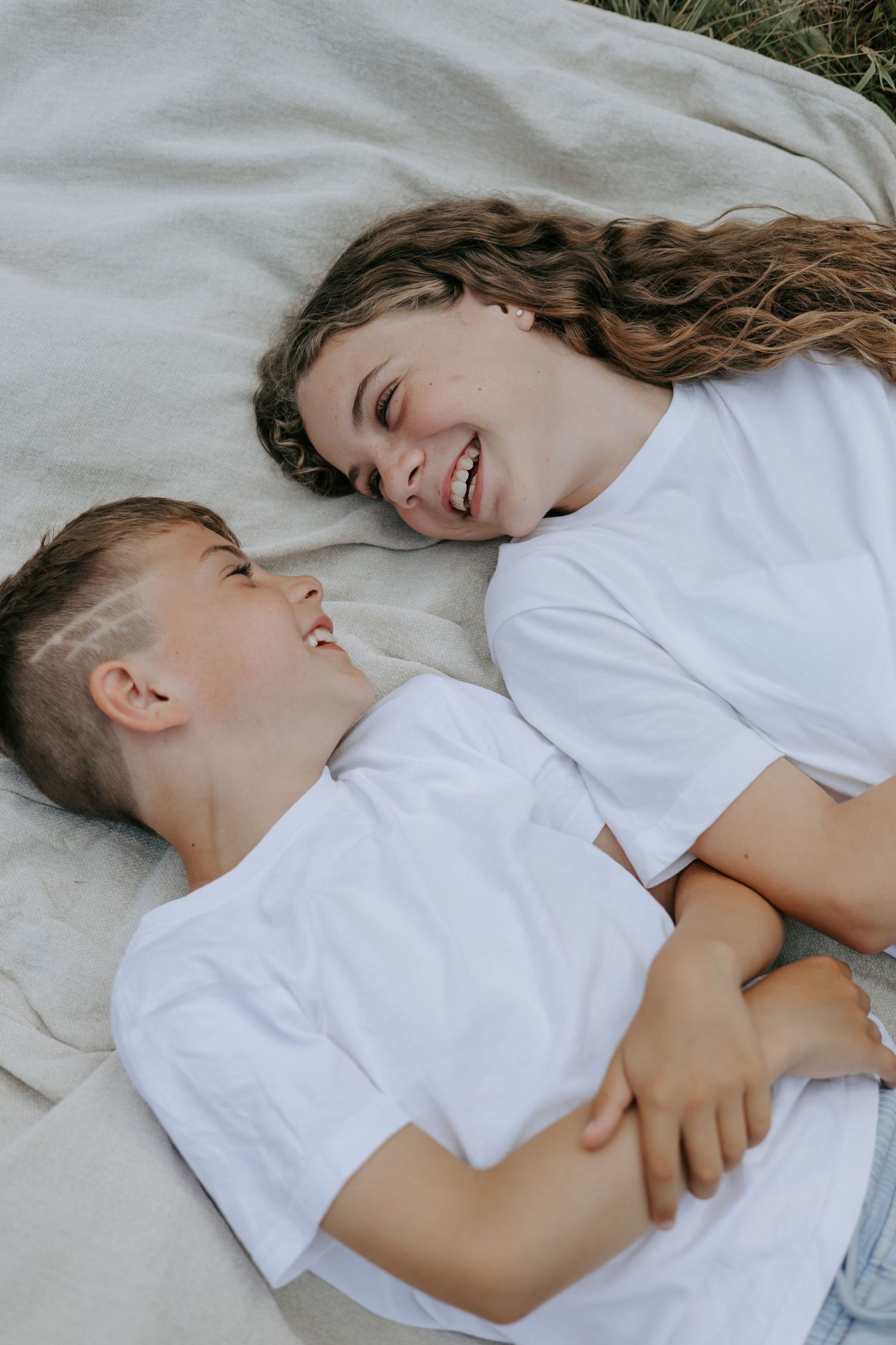 Two siblings lying on a blanket laughing together, wearing white t-shirts in a cozy outdoor setting.