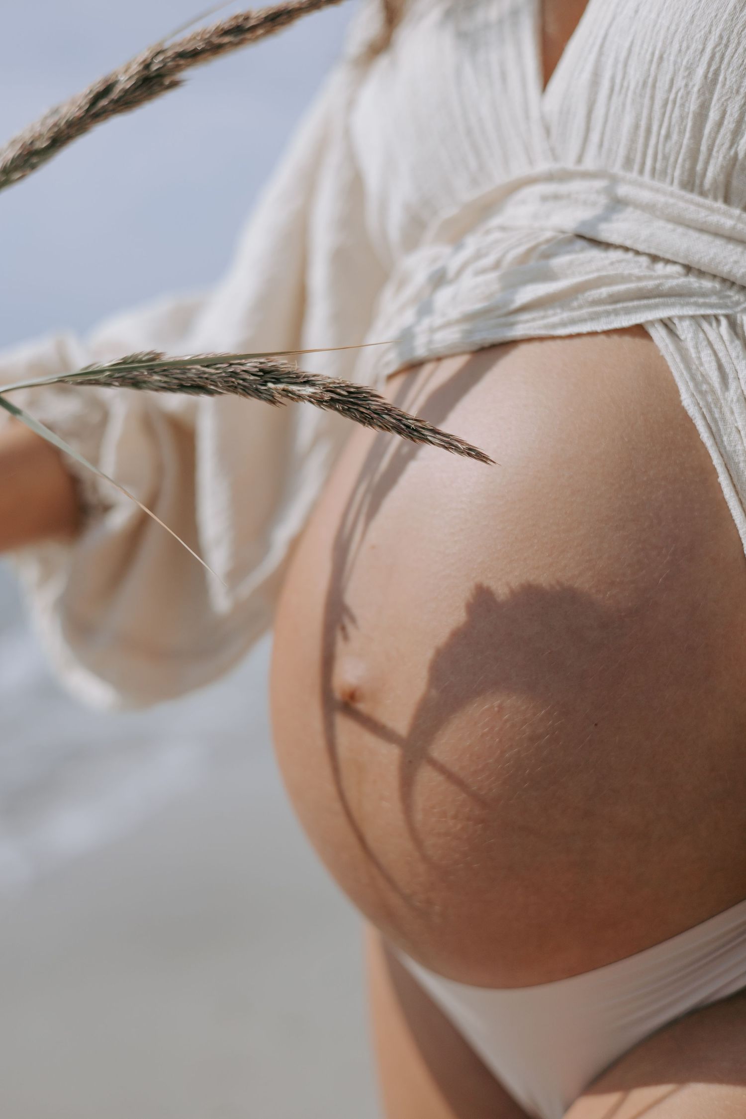 Close-up of pregnant belly with grass shadow, woman in white bikini and open linen robe at beach.