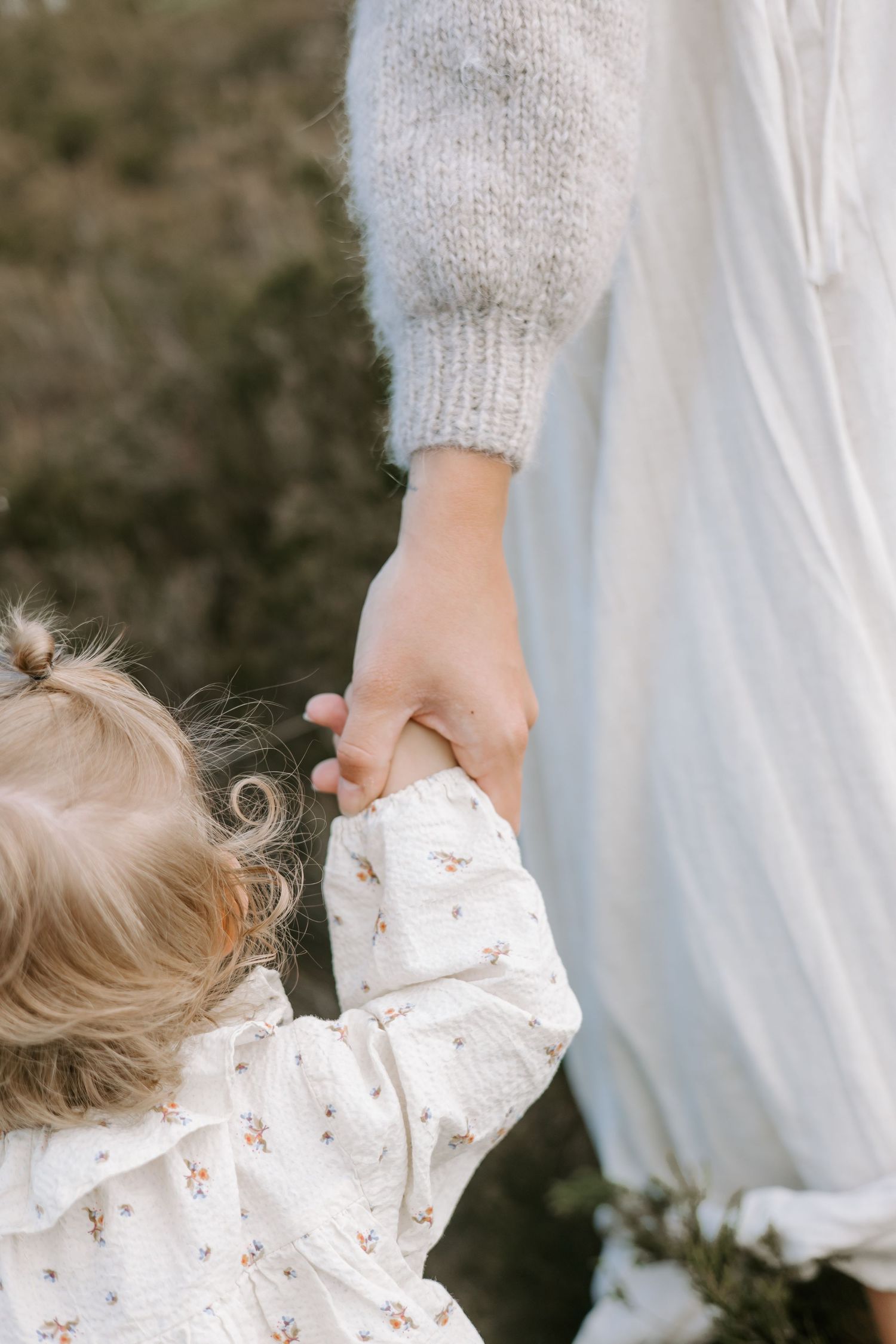 Young toddler holding mother's hand while walking outdoors in matching white outfits.