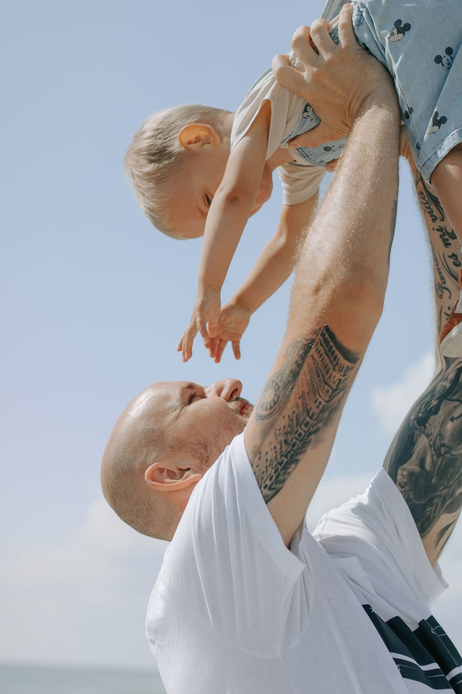 Tattooed father lifting smiling toddler high in the air against a blue sky.