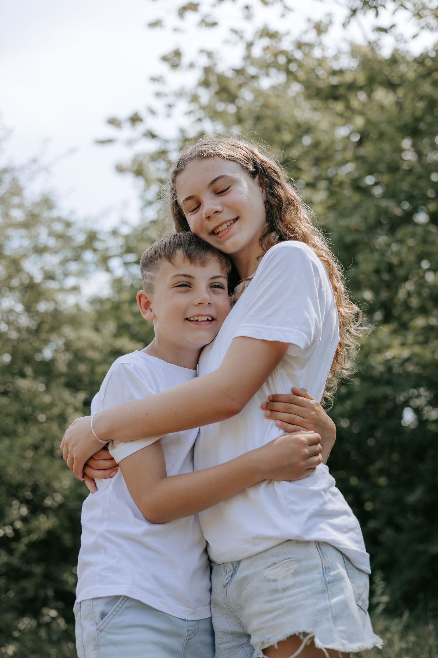 Smiling teenage girl hugging younger brother outdoors in white t-shirts with green trees behind them.