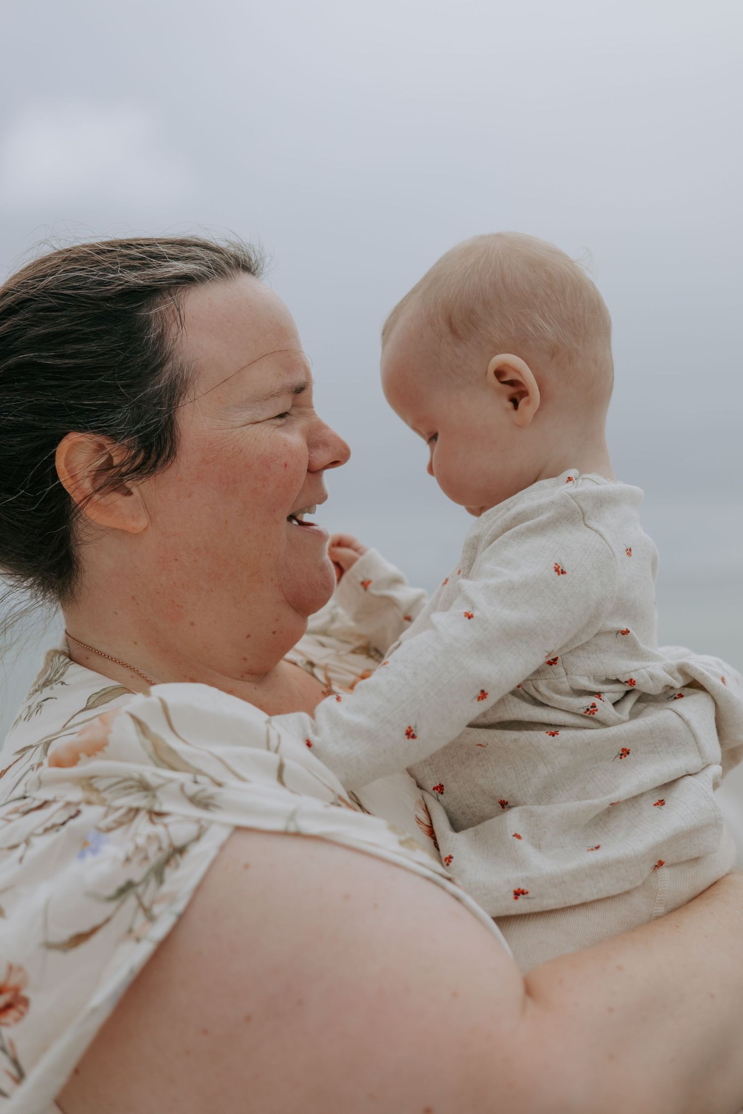 A joyful mother holding her smiling baby face-to-face outdoors on an overcast day.