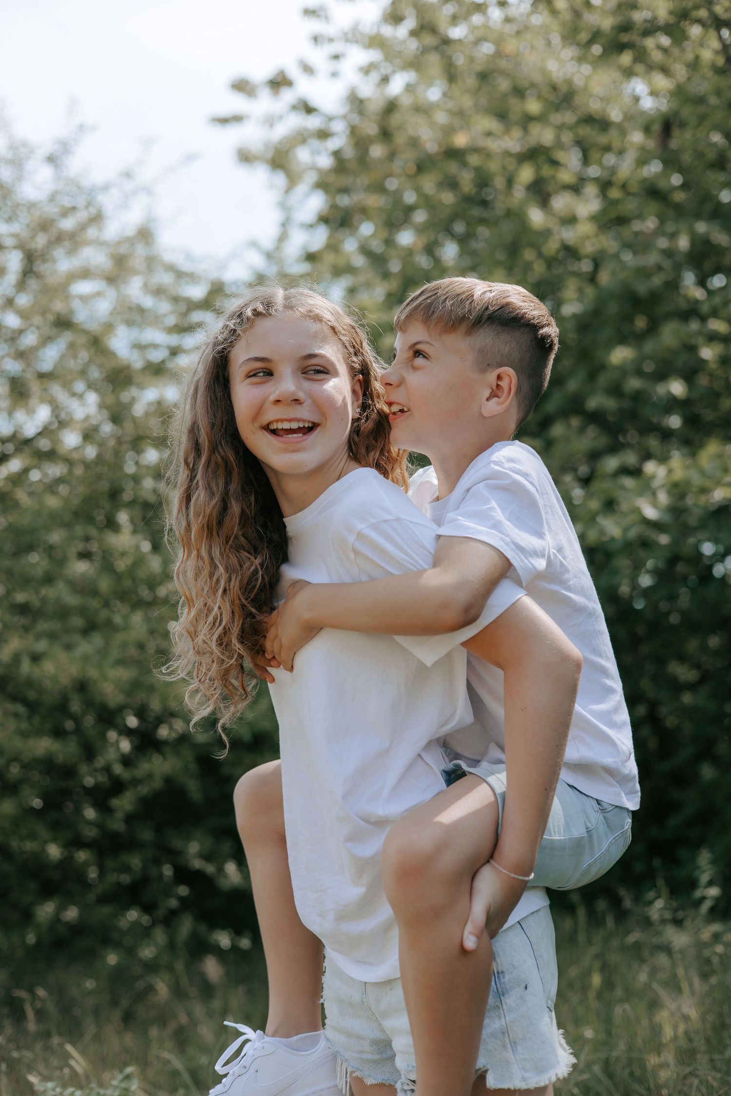 A laughing girl giving a boy a piggyback ride outdoors in a sunny green setting.