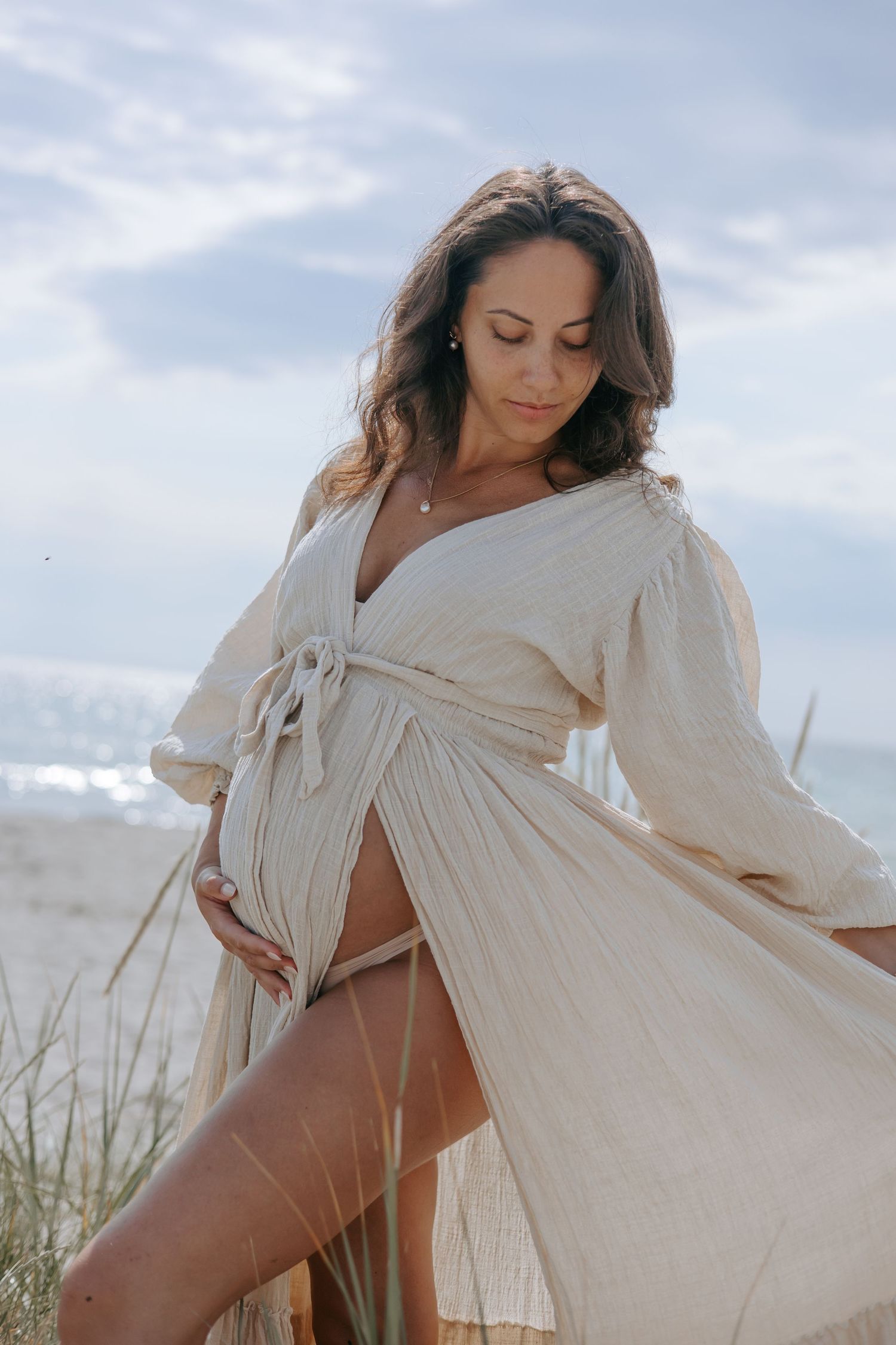 Pregnant woman in flowing beige wrap dress cradling baby bump on sandy beach with sea in background.