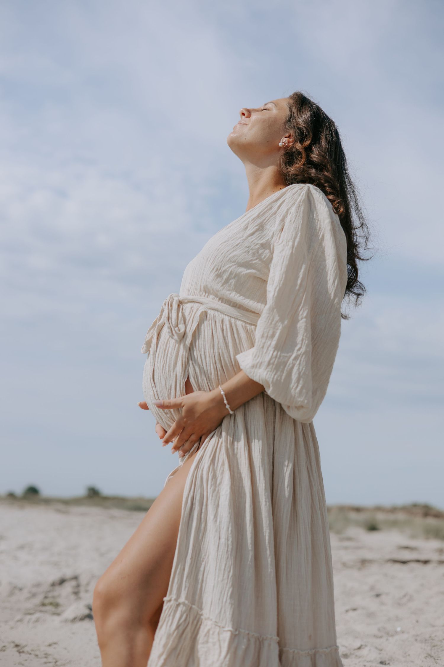 Pregnant woman in beige dress holding baby bump on sandy beach, head tilted back enjoying sunshine.