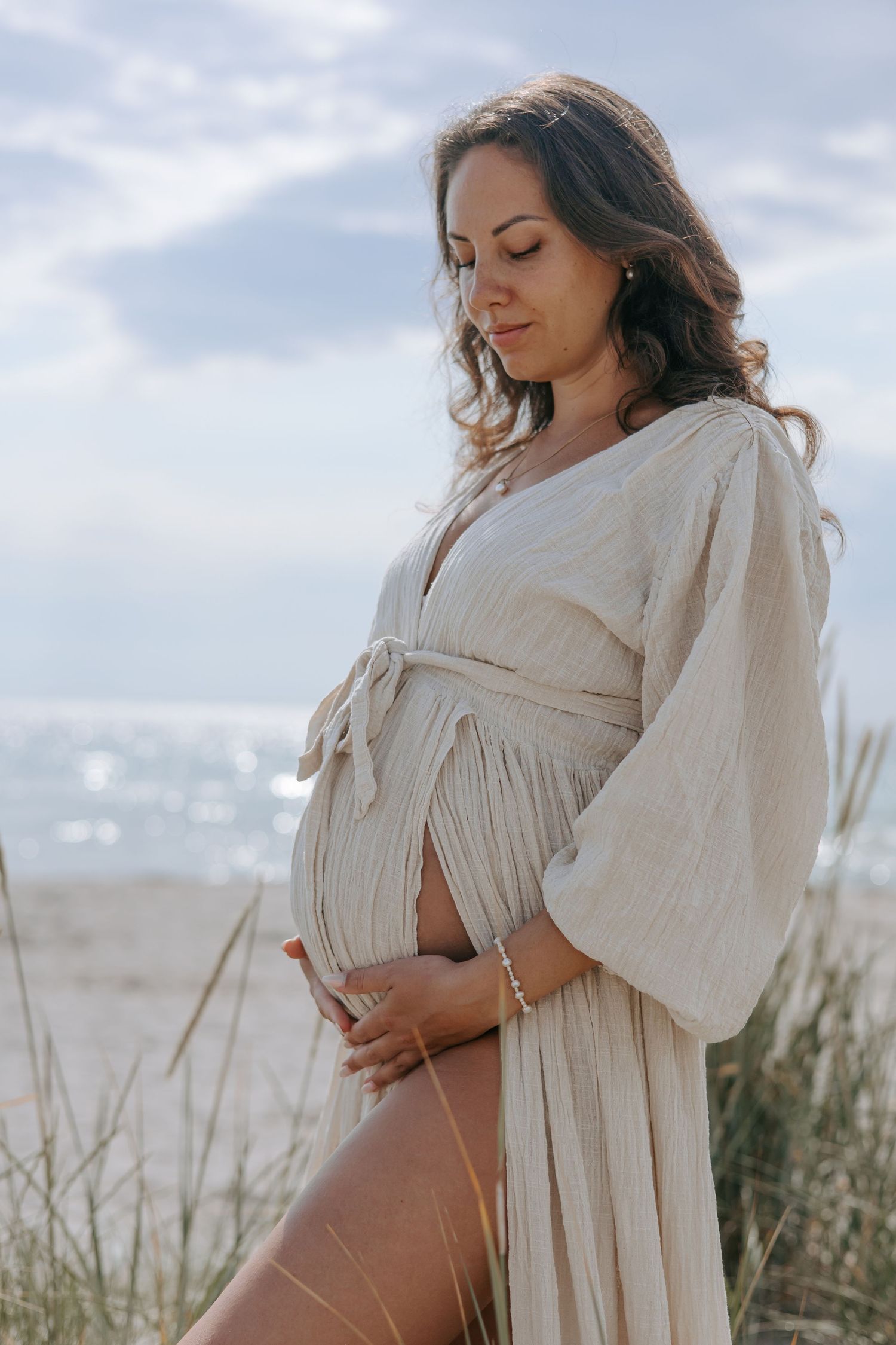 Pregnant woman in cream linen dress cradling baby bump on sandy beach with ocean behind.