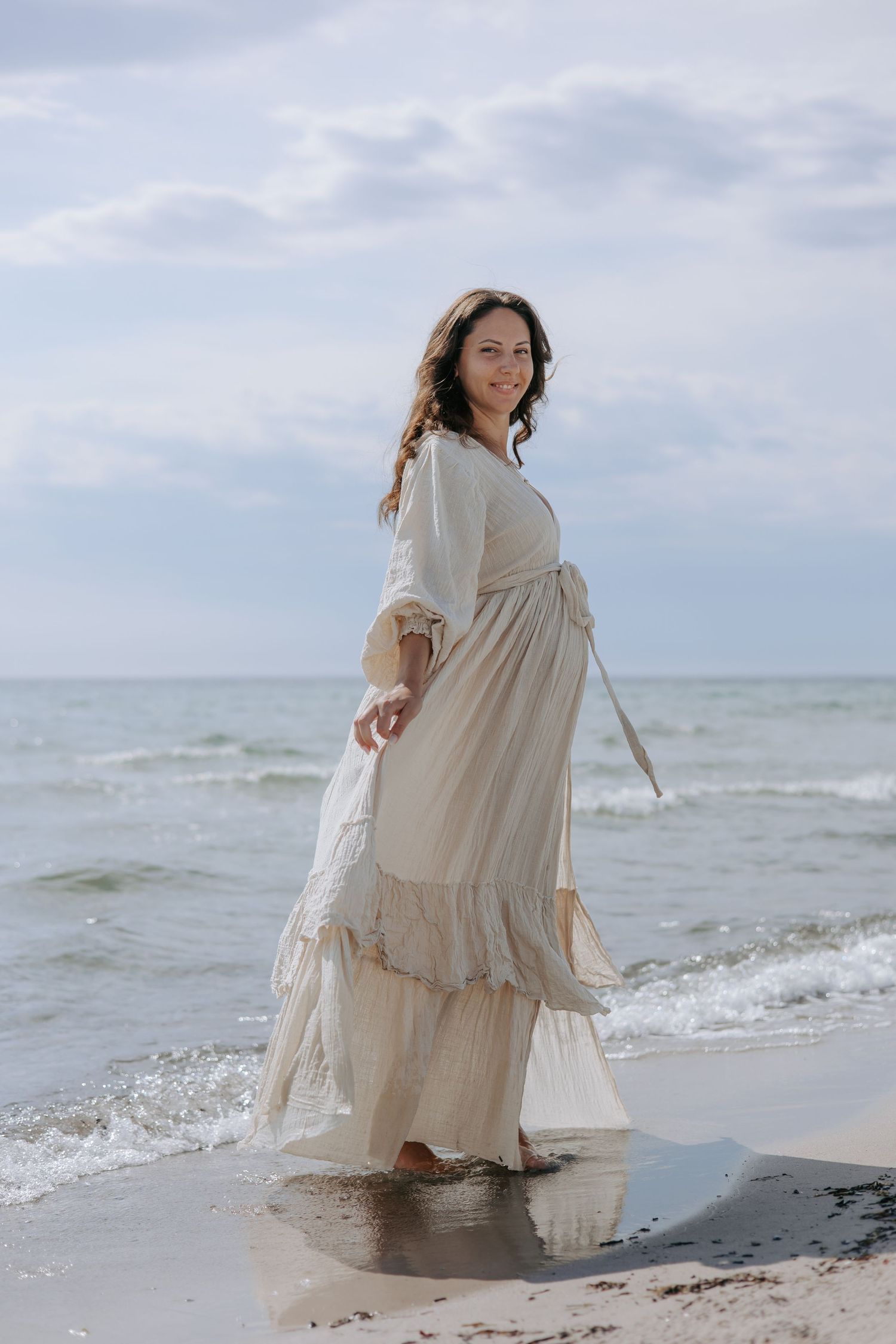 Smiling pregnant woman in white maxi dress walking along shoreline with ocean waves behind her.