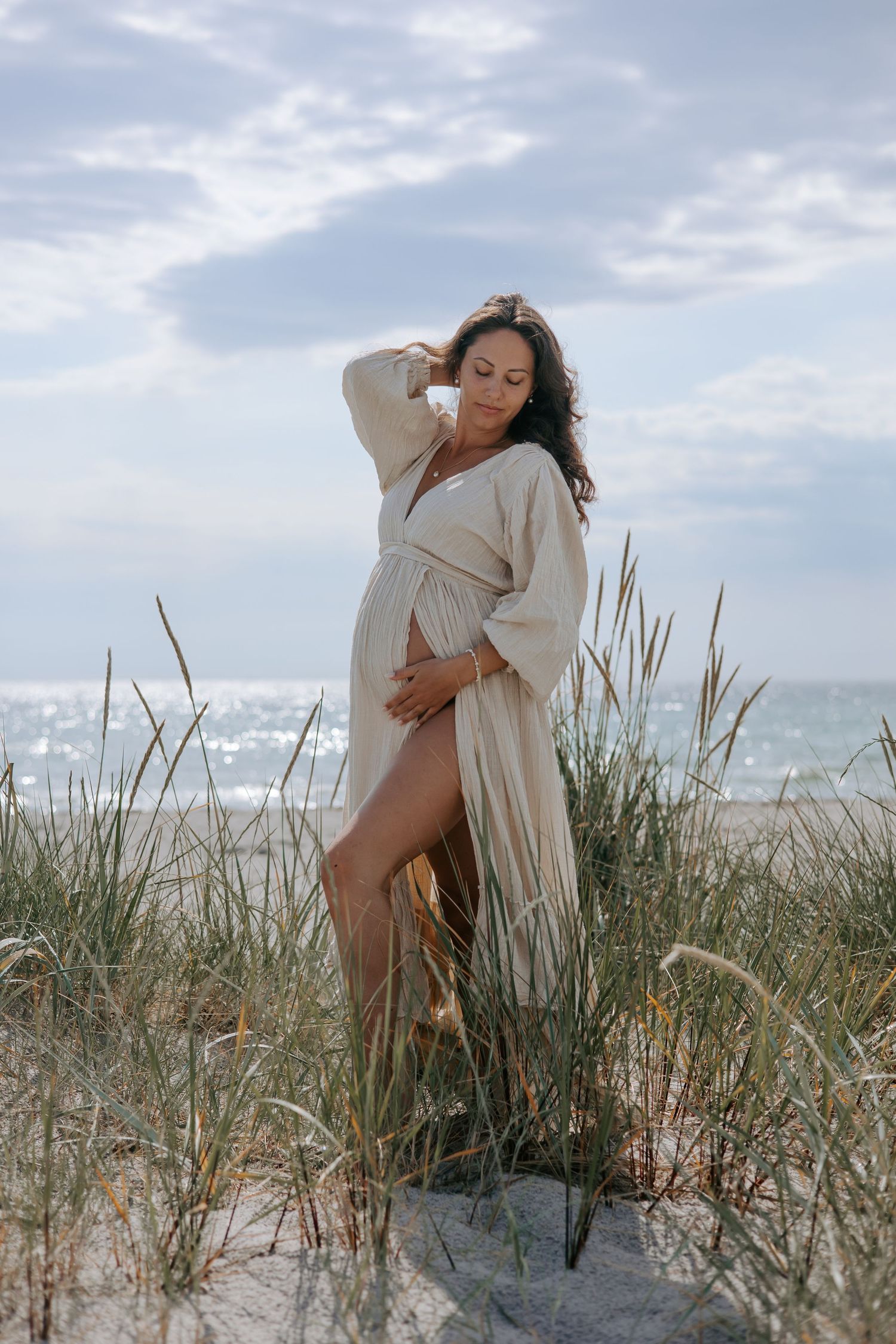 Pregnant woman in a flowing beige linen dress standing in sandy beach dunes, gently holding her belly.