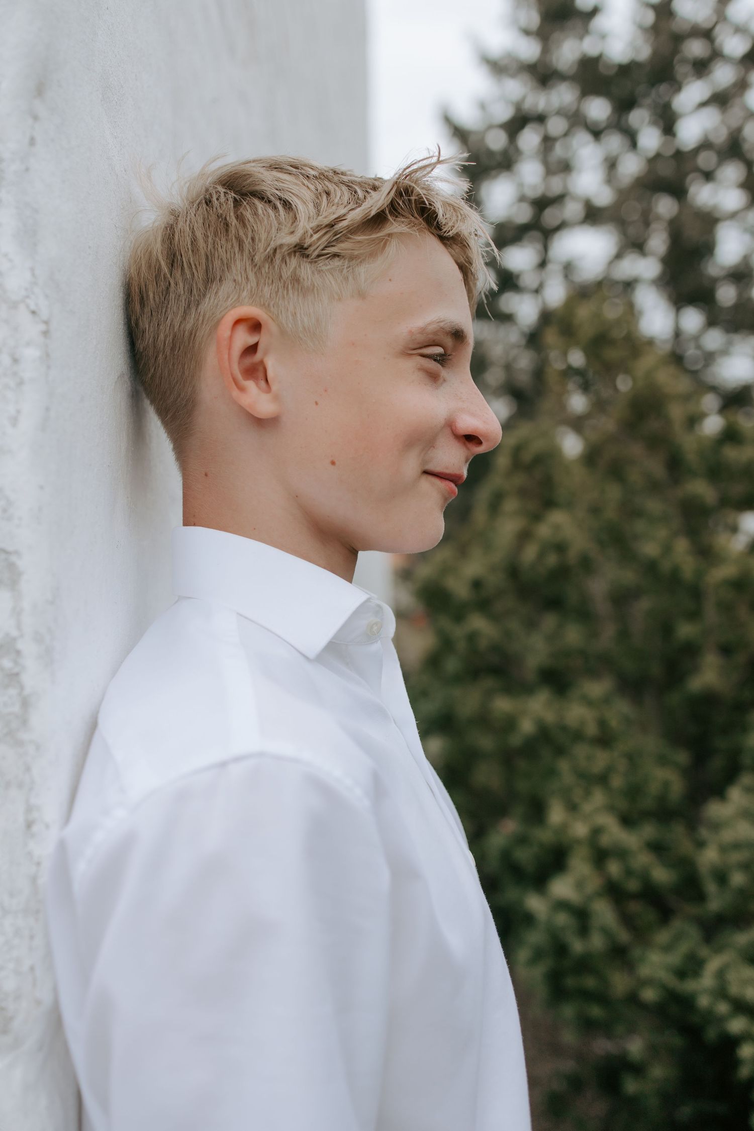 Young blonde teen boy in white shirt leaning against white wall outdoors.