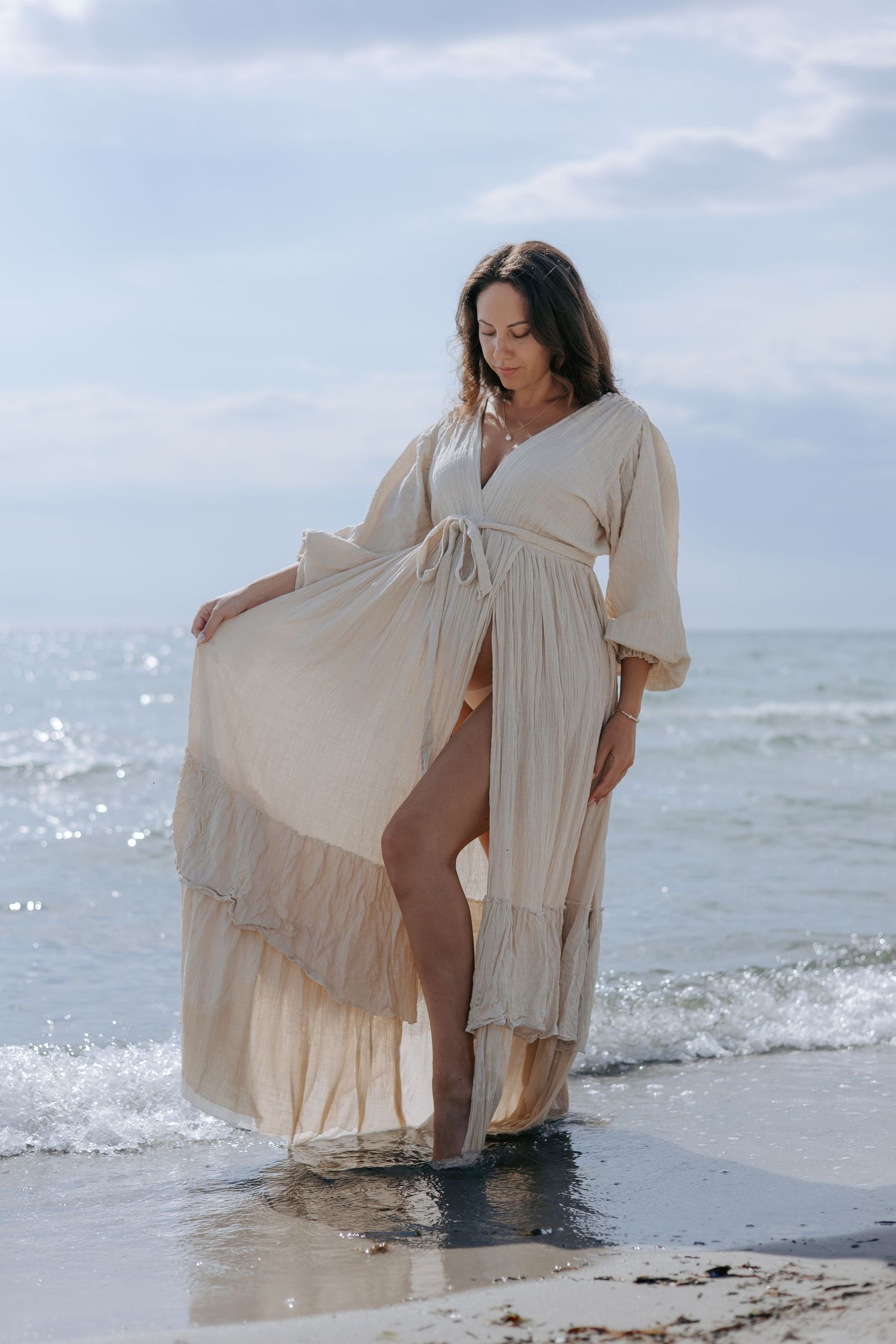 Pregnant woman in flowing beige dress standing at ocean shoreline with waves and cloudy sky.