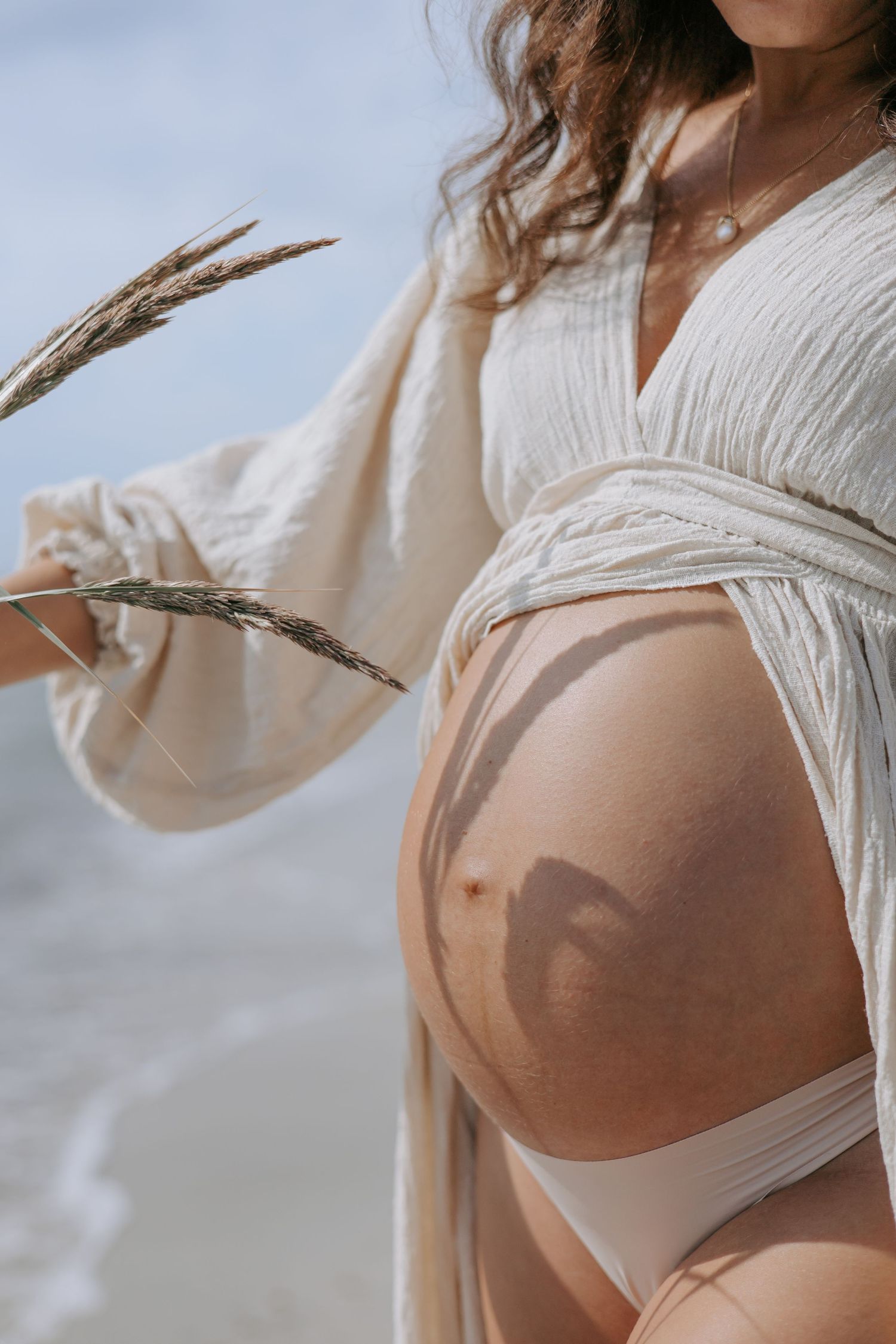 Pregnant woman in white outfit holding dried grass near ocean beach.
