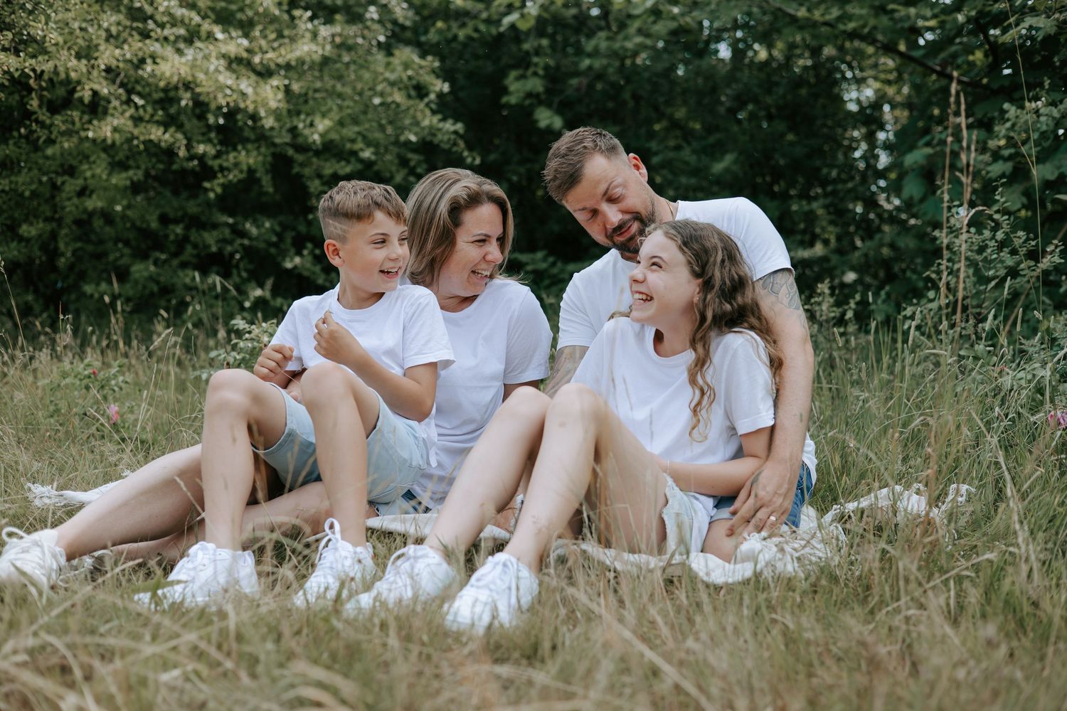 Happy family of four sitting together on a blanket in a green meadow, all wearing white t-shirts.
