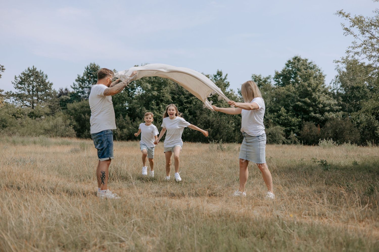 Family of four playing with a blanket in an open grassy field on a cloudy day, wearing casual outfits.