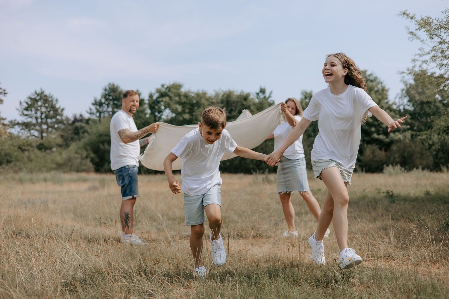 Joyful family of four running and playing together in a sunny field, wearing matching white outfits.