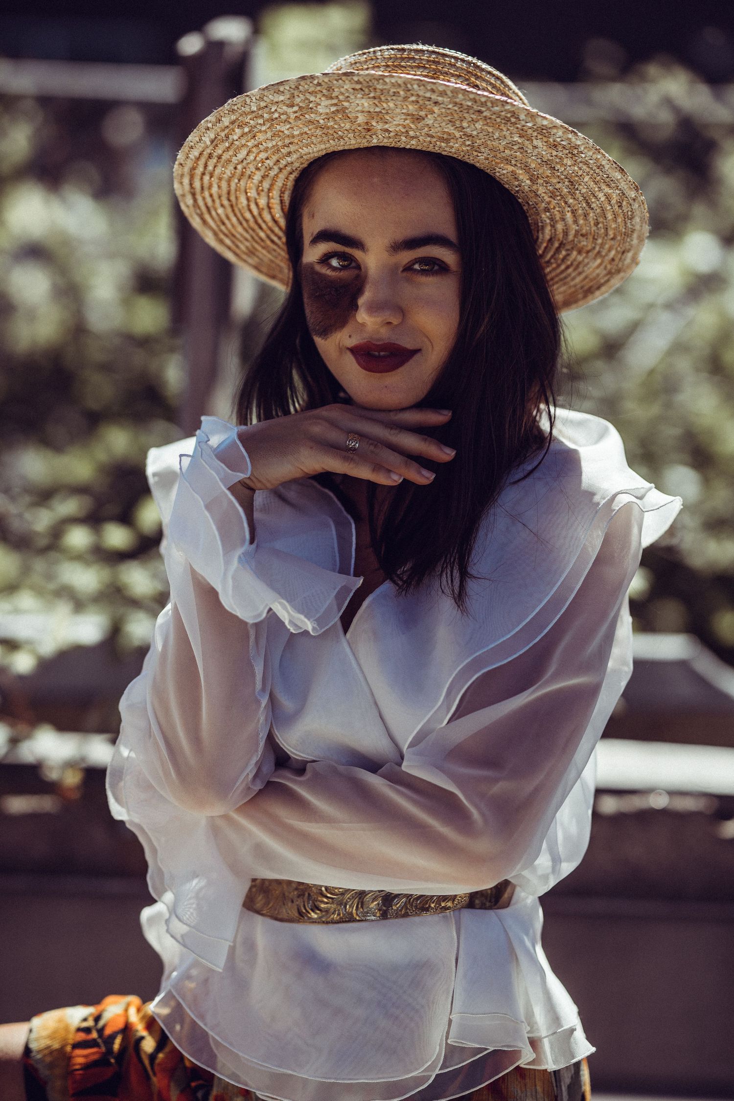 Young woman in straw hat and white ruffled blouse with gold belt poses outdoors in dappled sunlight.