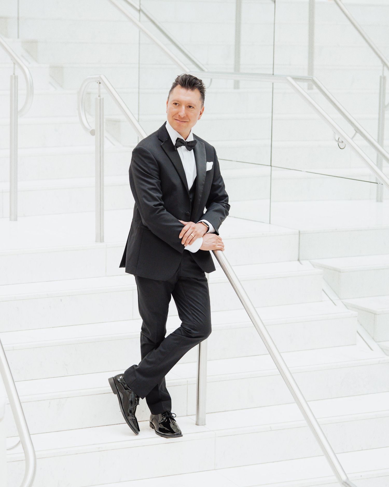 Man in black tuxedo with bow tie posing on white marble stairs, leaning on metal railing.