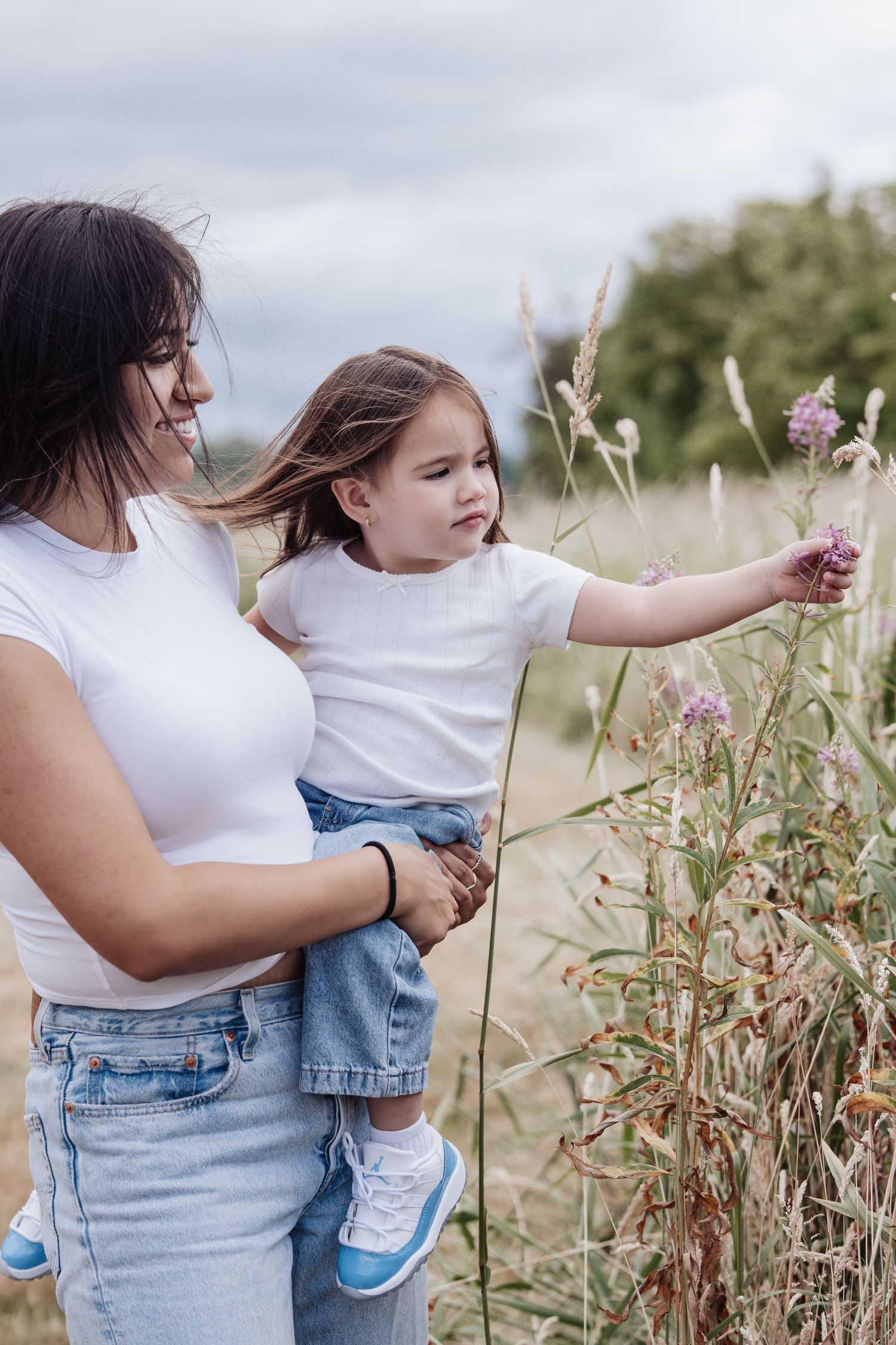 A mother holds her young daughter while she reaches out to touch wildflowers in a rustic field.