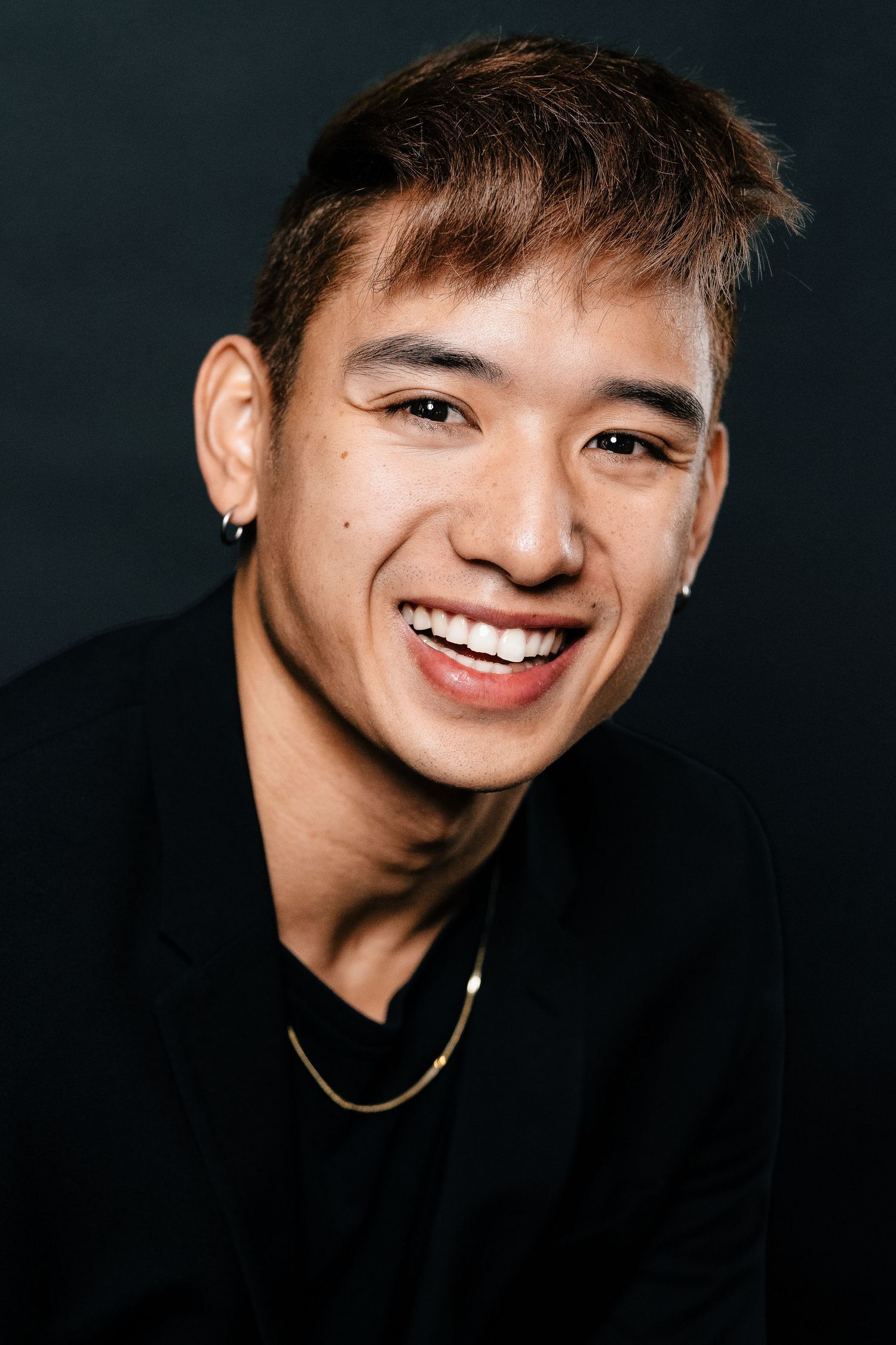 Smiling young Asian man with ear stud and gold chain necklace wearing black jacket against dark background.