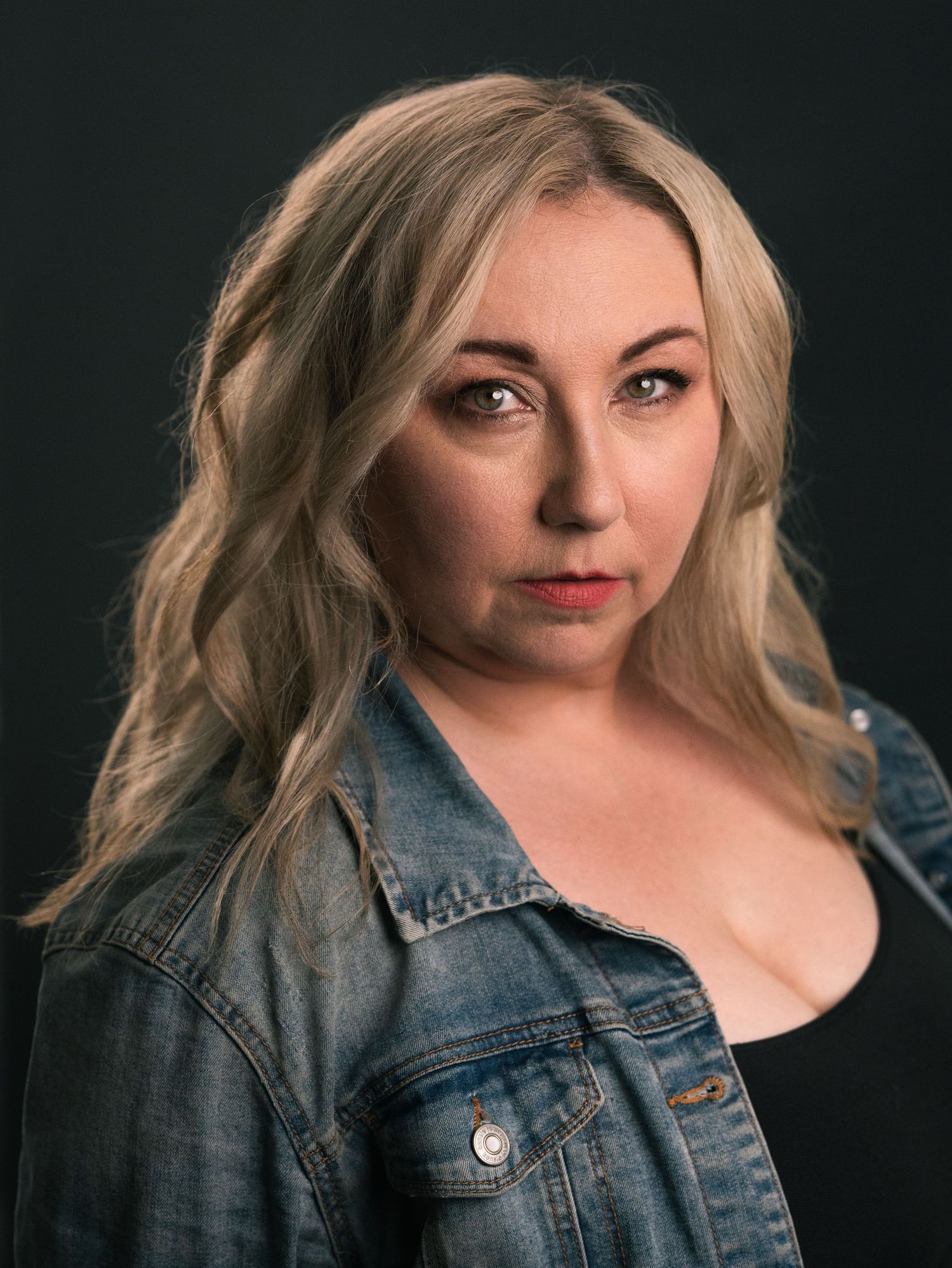 Blonde woman in denim jacket posing against dark background for professional headshot portrait.