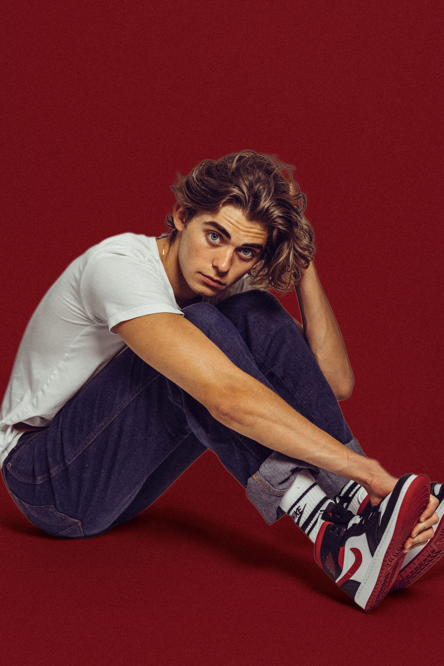 Young man with curly hair sitting on red background wearing white tee, jeans, and red Nike Air Jordan sneakers.