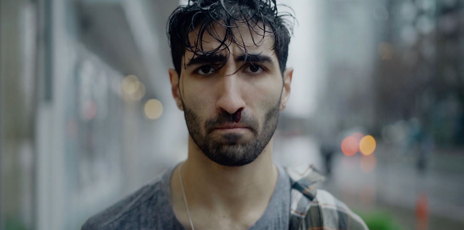 Close-up portrait of a young man with dark hair and beard standing on a rainy urban street.