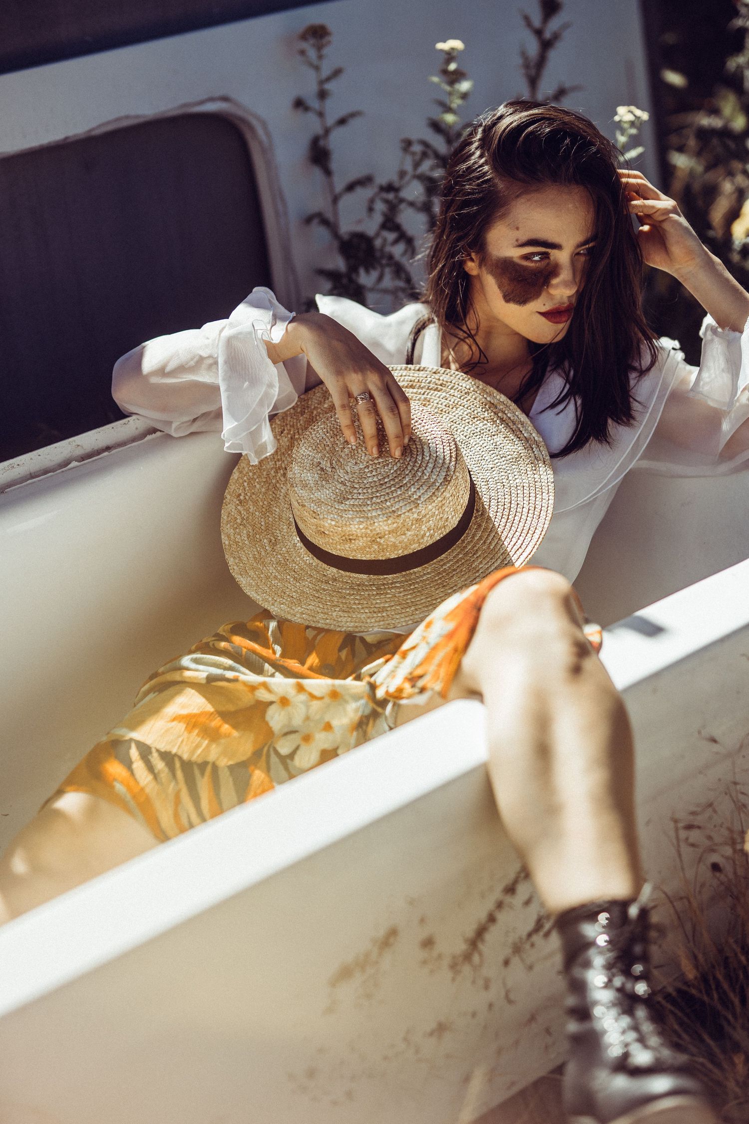 Woman in white blouse and floral skirt holding straw hat while reclining in vintage outdoor bathtub.