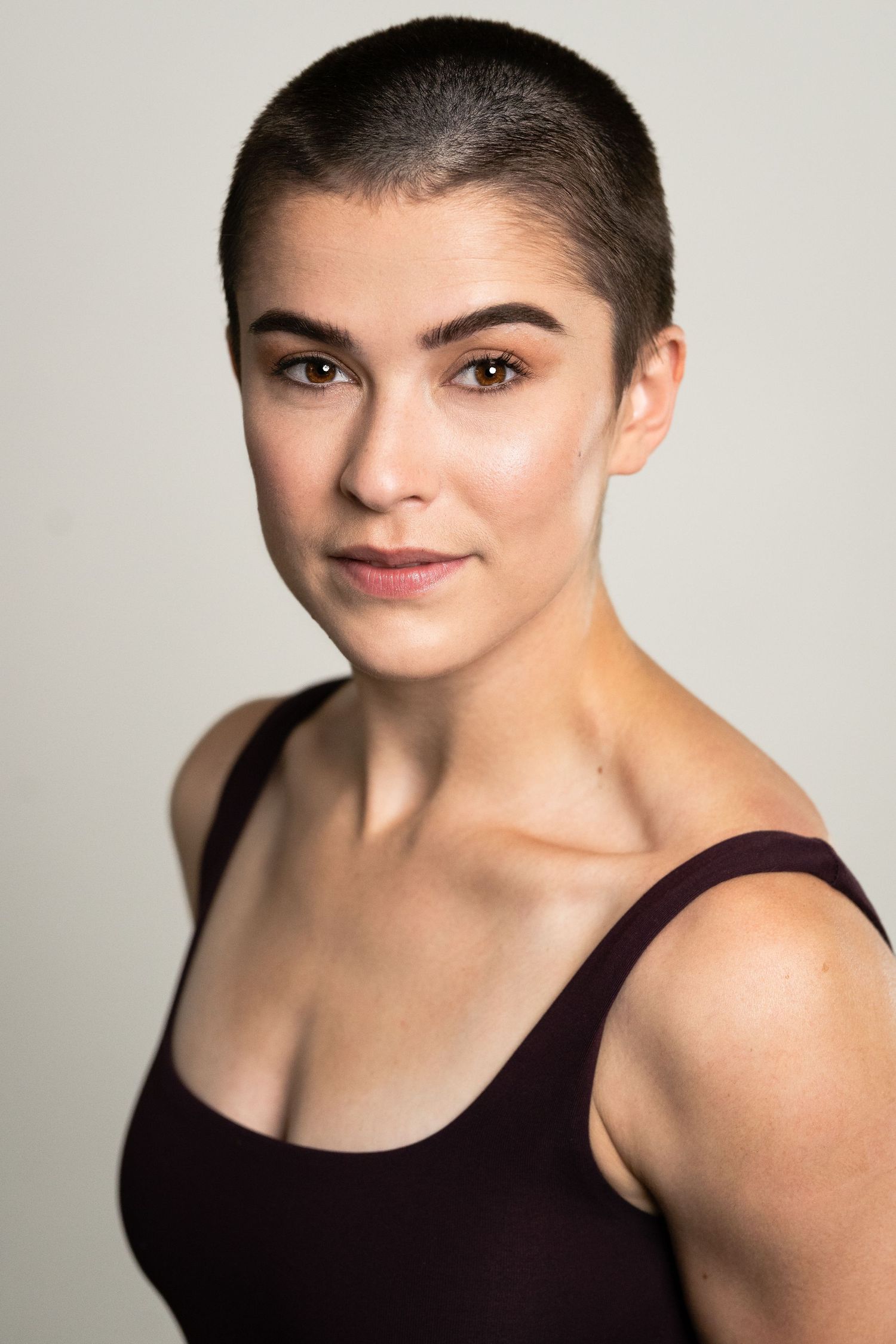 Young woman with buzz cut hair wearing a dark maroon tank top poses for a professional headshot portrait.