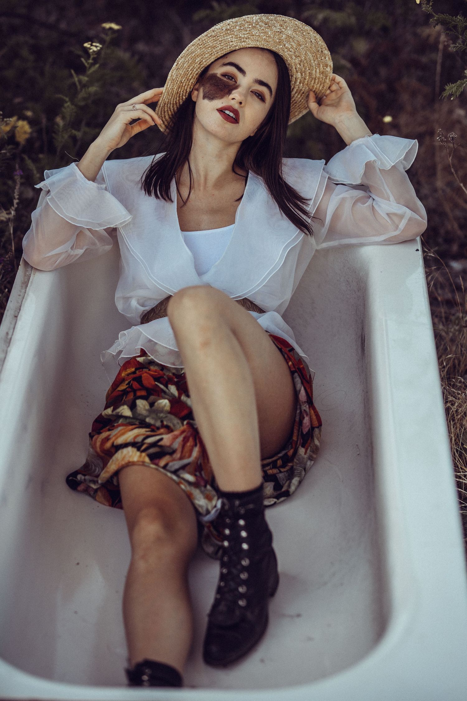Young woman in straw hat, sheer blouse, floral skirt, and black boots posing in a vintage bathtub outdoors.