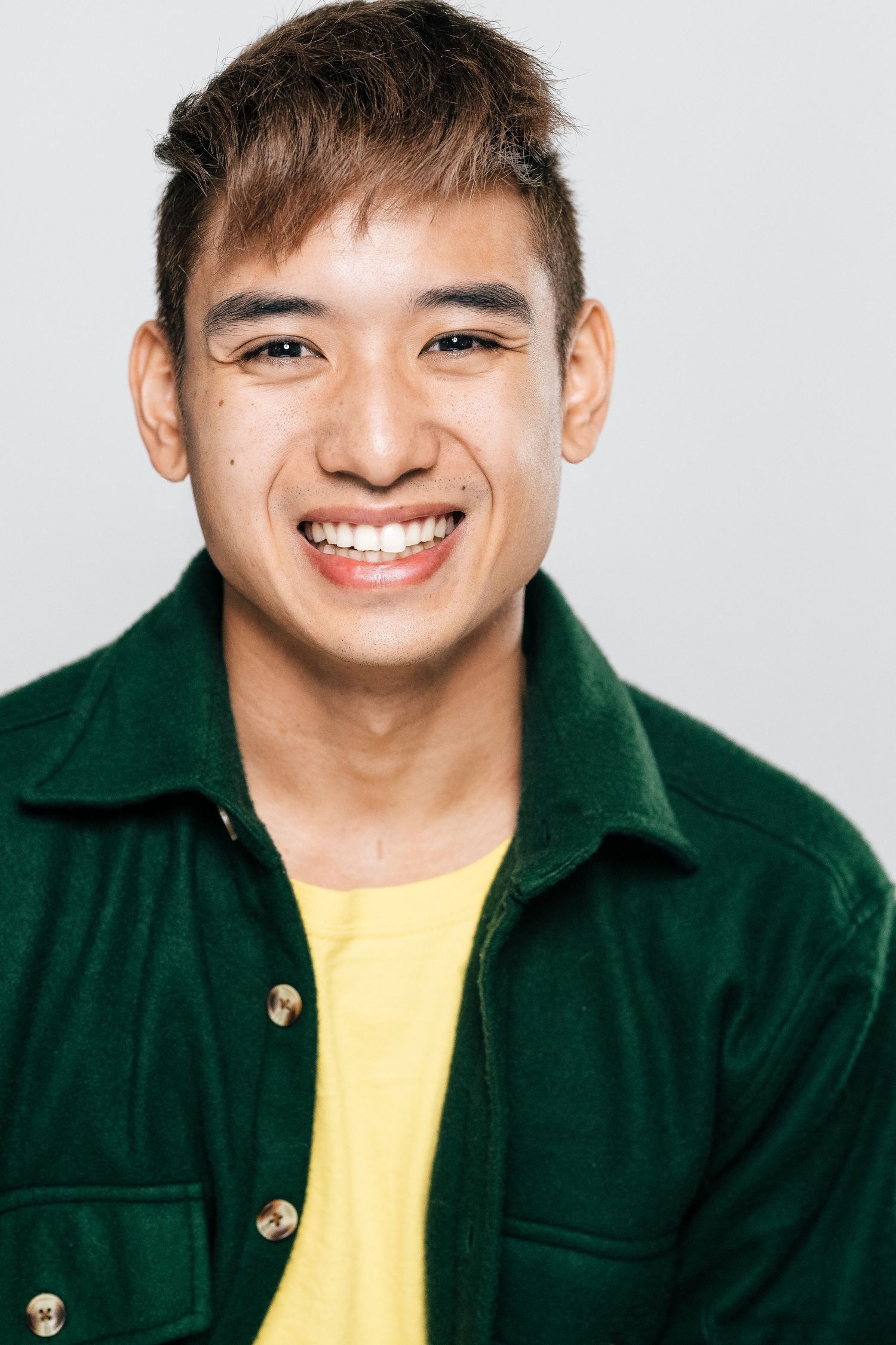 Smiling young man wearing a green jacket over a yellow shirt against a light gray background.