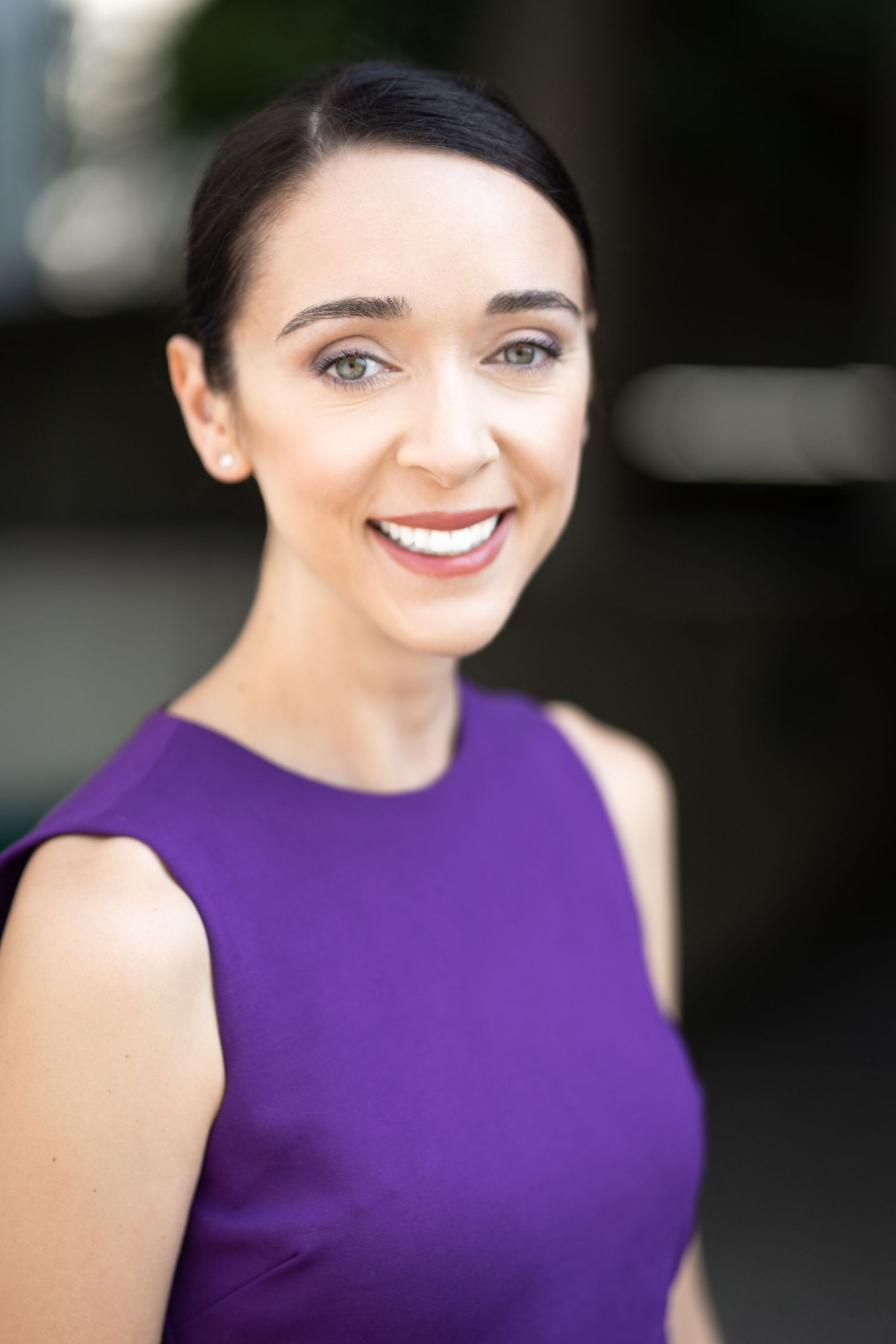 Smiling woman with dark hair in a purple sleeveless dress poses outdoors for a professional headshot photo.