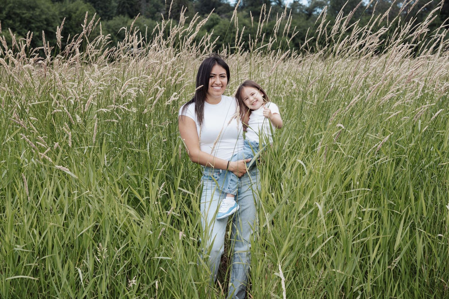 A mother and daughter smiling together in a lush green field of tall grass, both wearing jeans and white tops.
