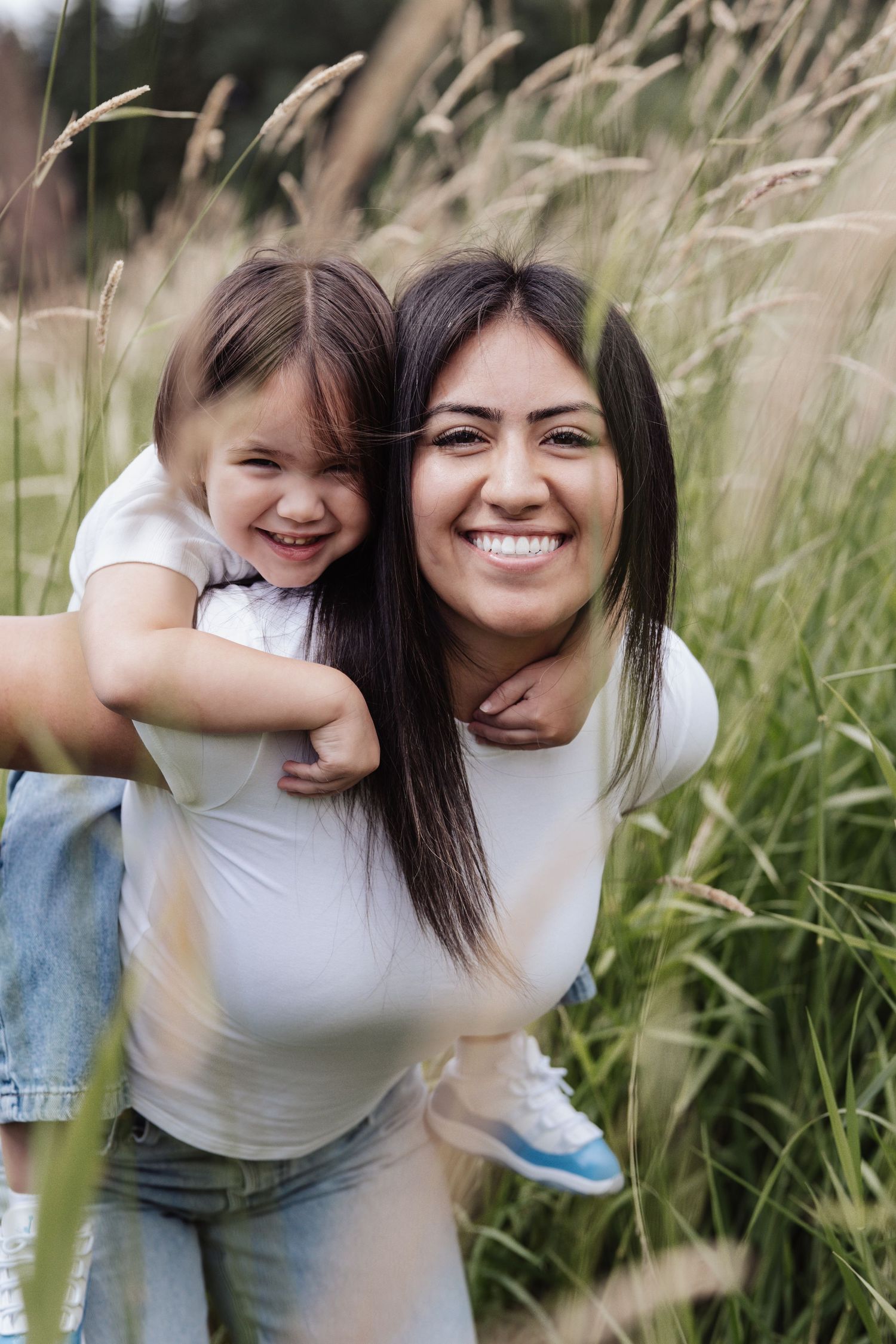 A smiling woman gives a happy young girl a piggyback ride through tall grass in an outdoor field.