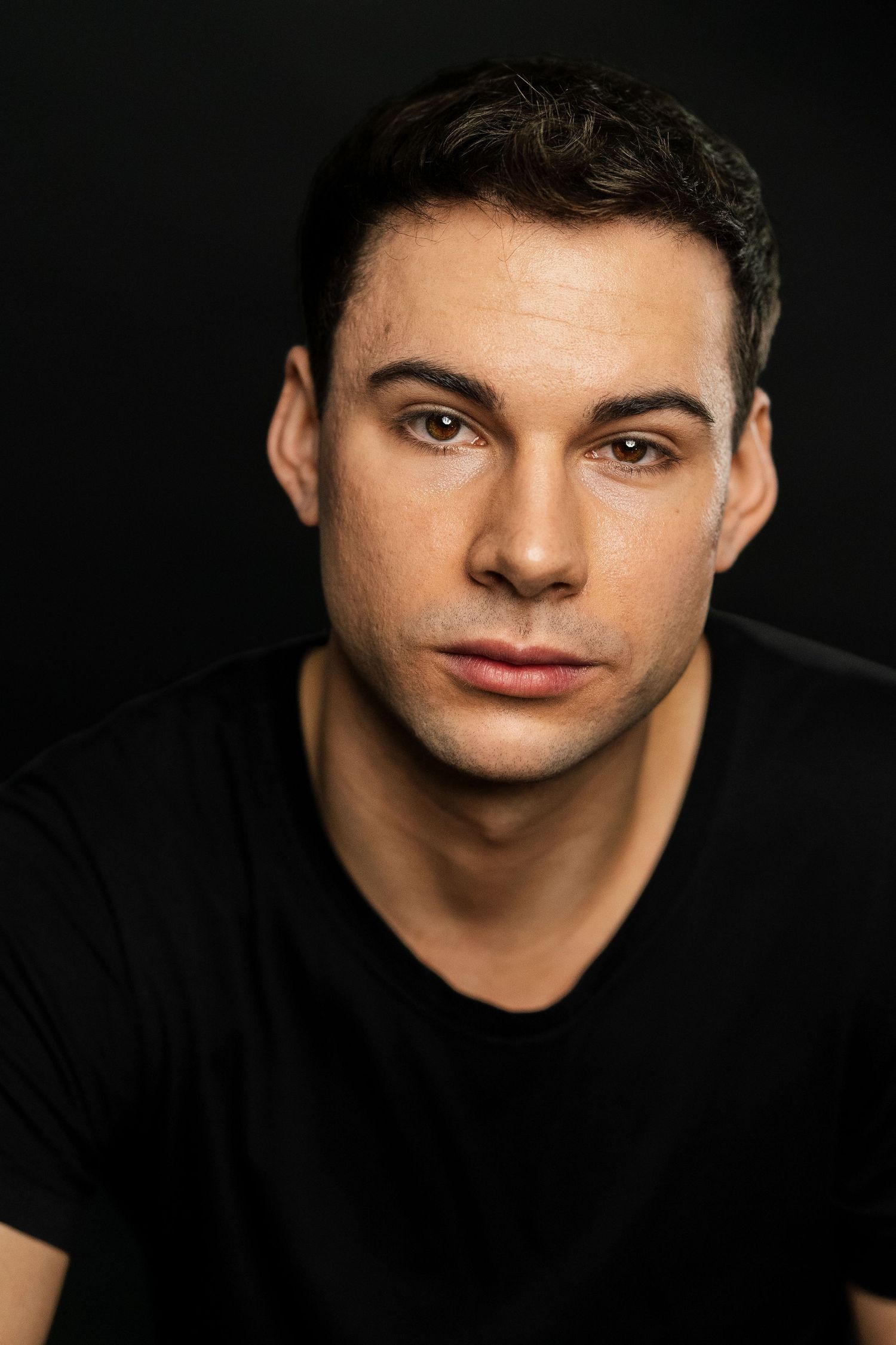 Young man with dark hair wearing a black t-shirt poses against a dark background in a professional headshot.