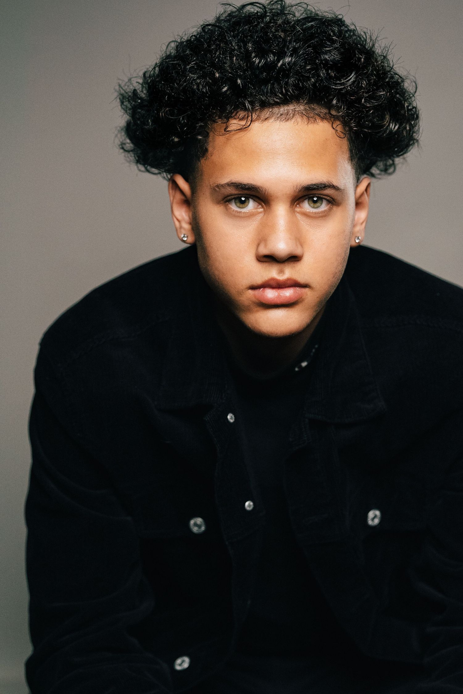 Young man with curly black hair and small earrings wearing a black jacket, posing against a gray background.