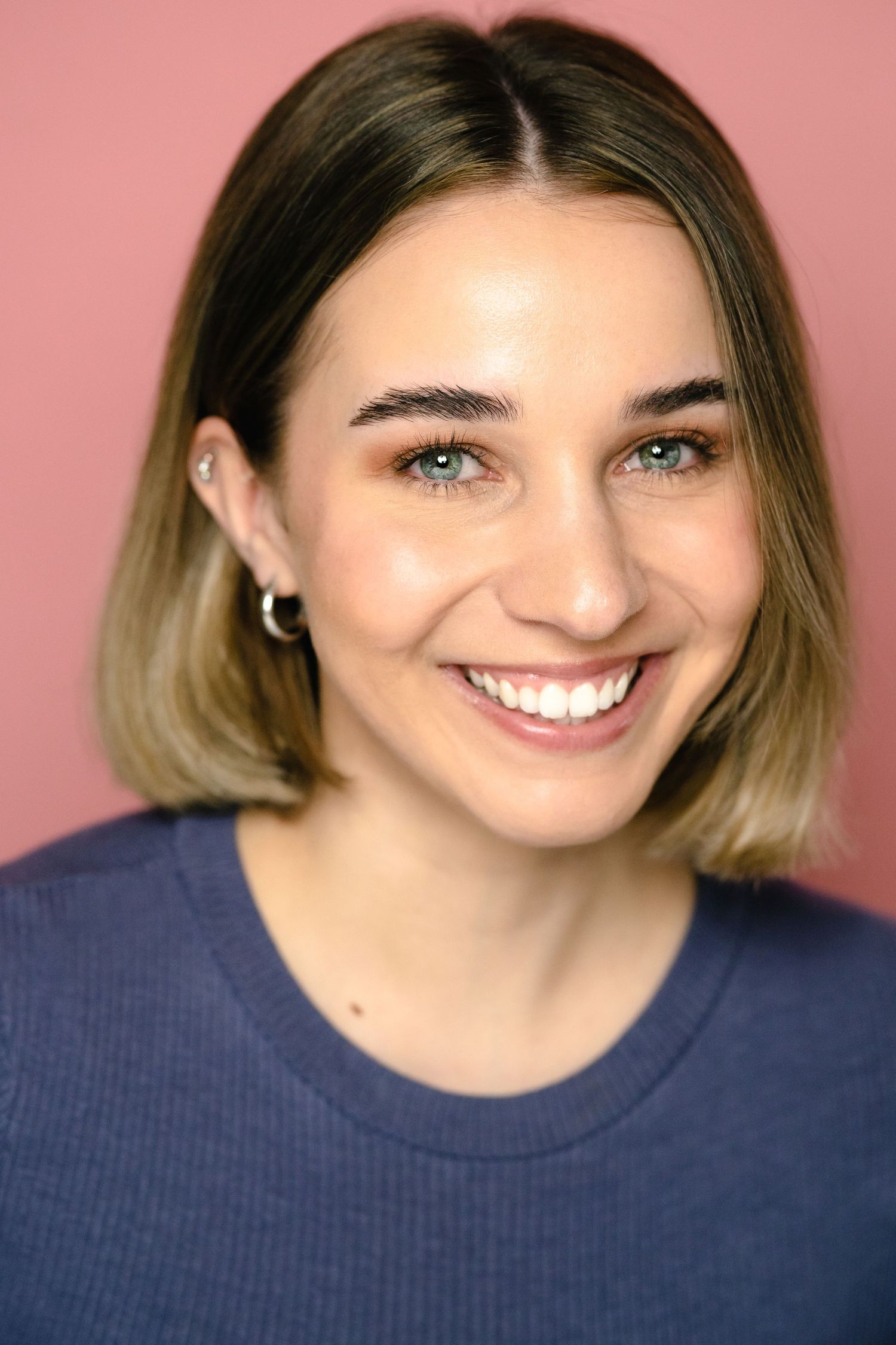 Smiling young woman with short blonde hair and blue eyes wearing a navy top against a pink background.