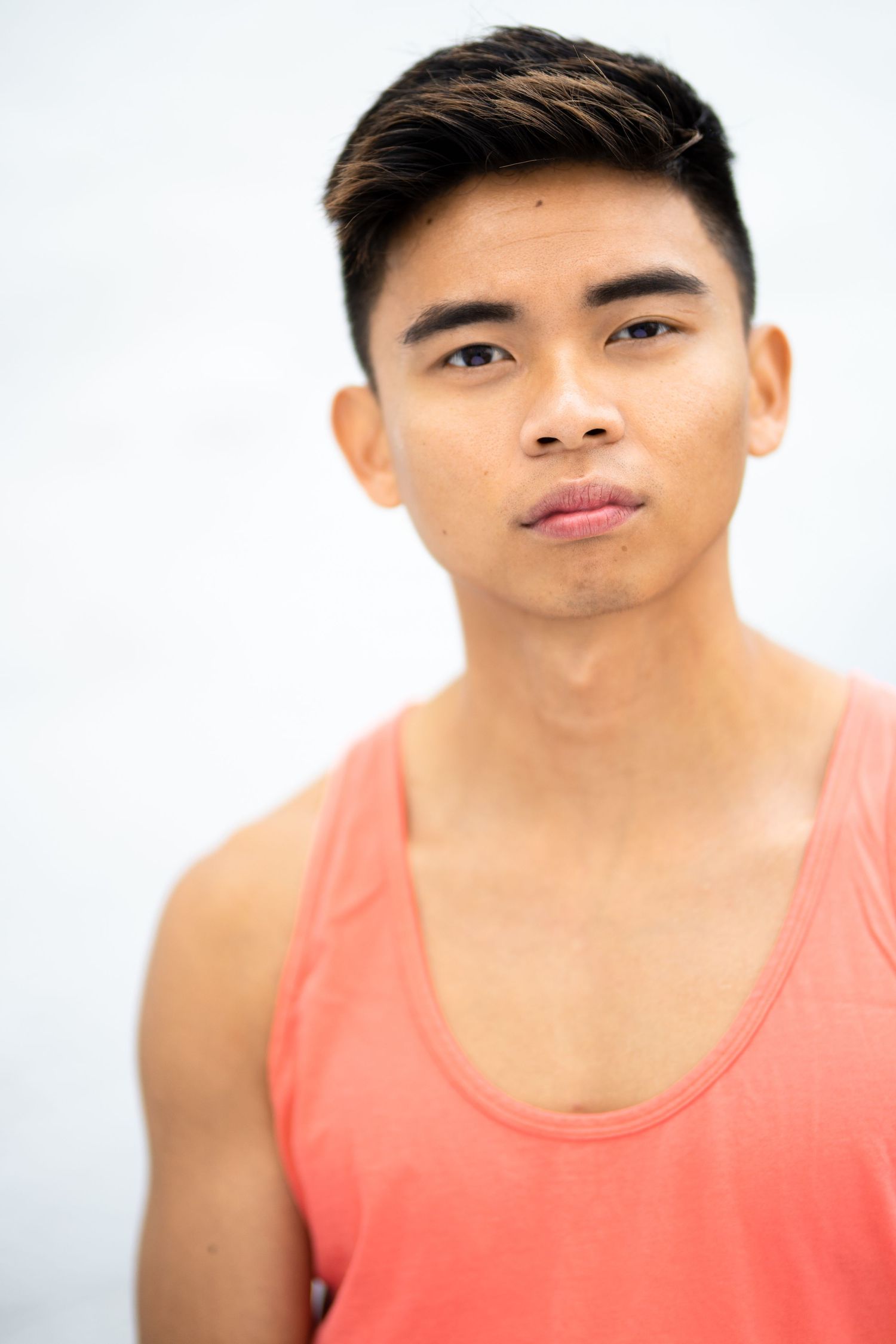 Young Asian man in a coral tank top posing against a light gray background with a serious expression.