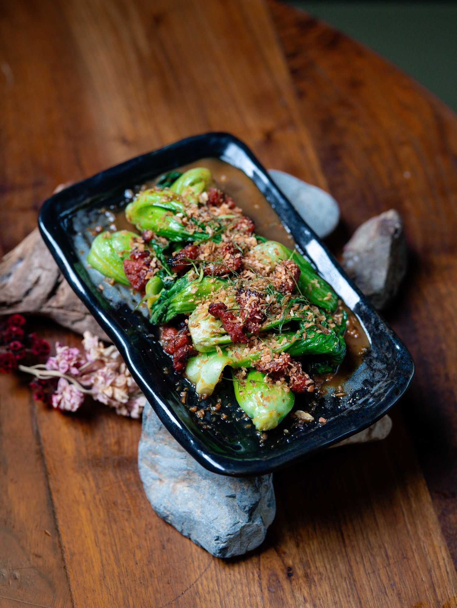 Braised bok choy topped with spiced meat sauce served on a black rectangular plate resting on stones.