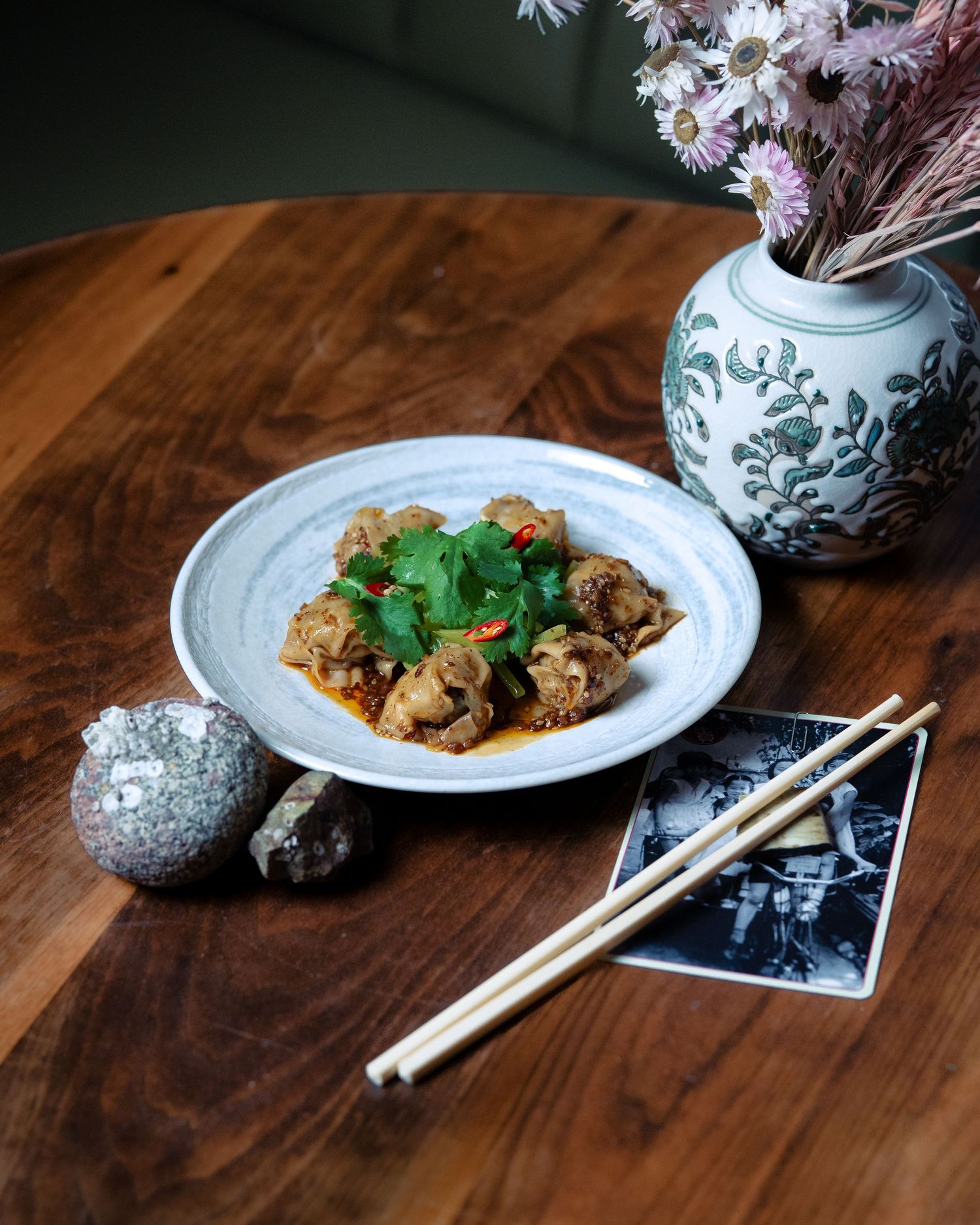 Asian dish with herbs and chili on ceramic plate, chopsticks, floral vase on wooden table.